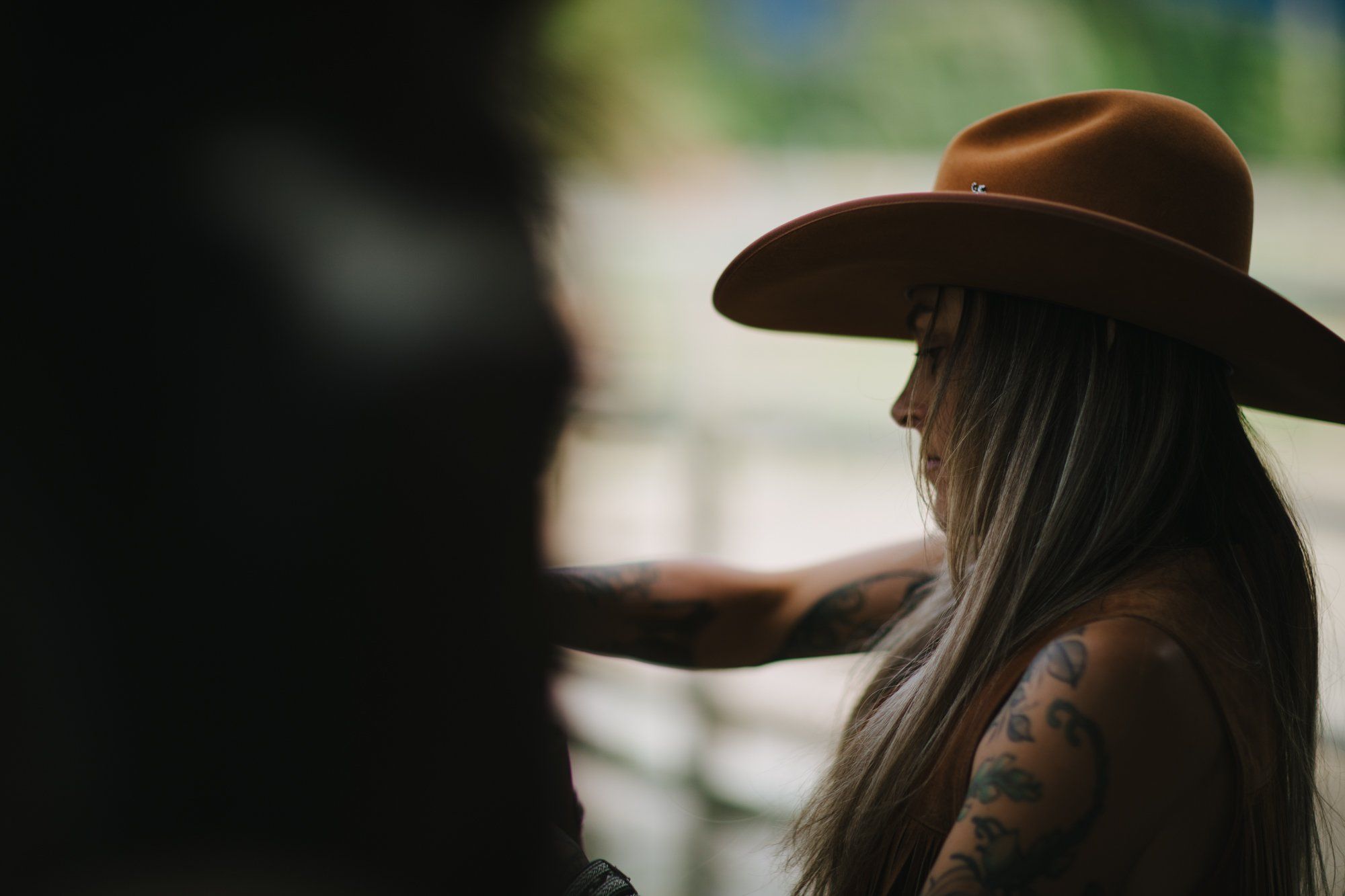 Woman in brown cowboy hat with long dark hair and arm tattoos, backlit against blurred green landscape