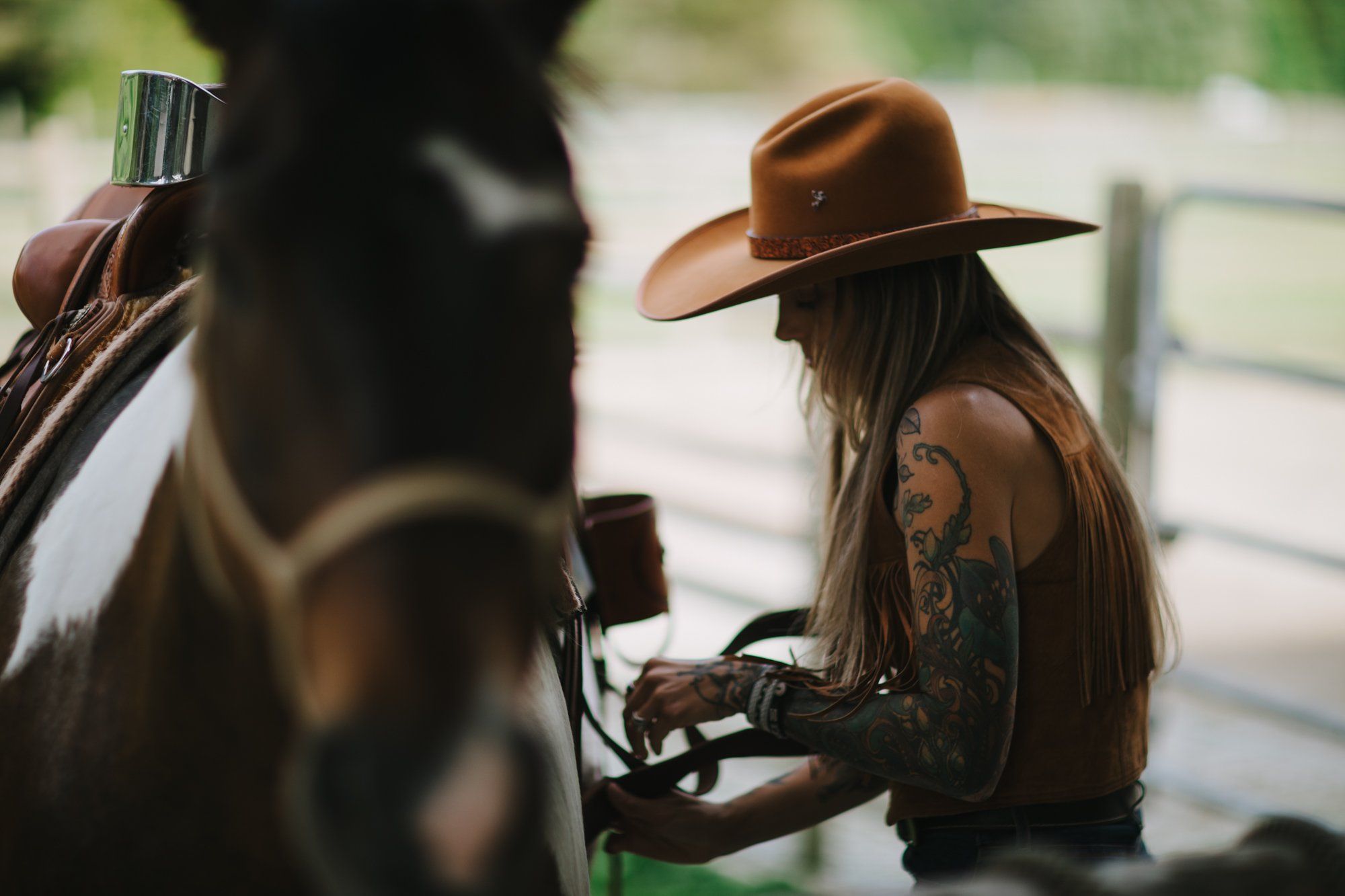 Tattooed cowgirl in brown hat saddling horse in golden afternoon light, focus soft on her determined expression and weathered leather gear
