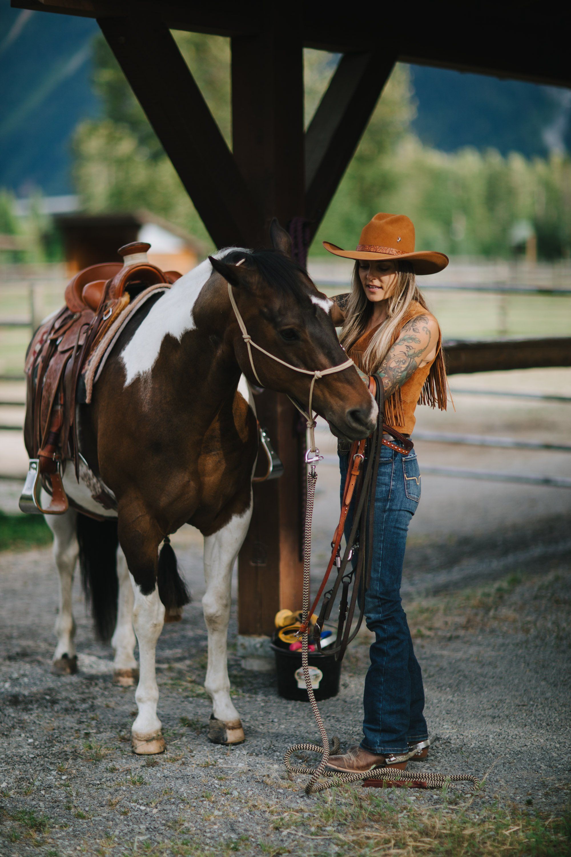 Young girl in cowboy hat holds brown and white horse under wooden structure, wearing denim and fringe jacket