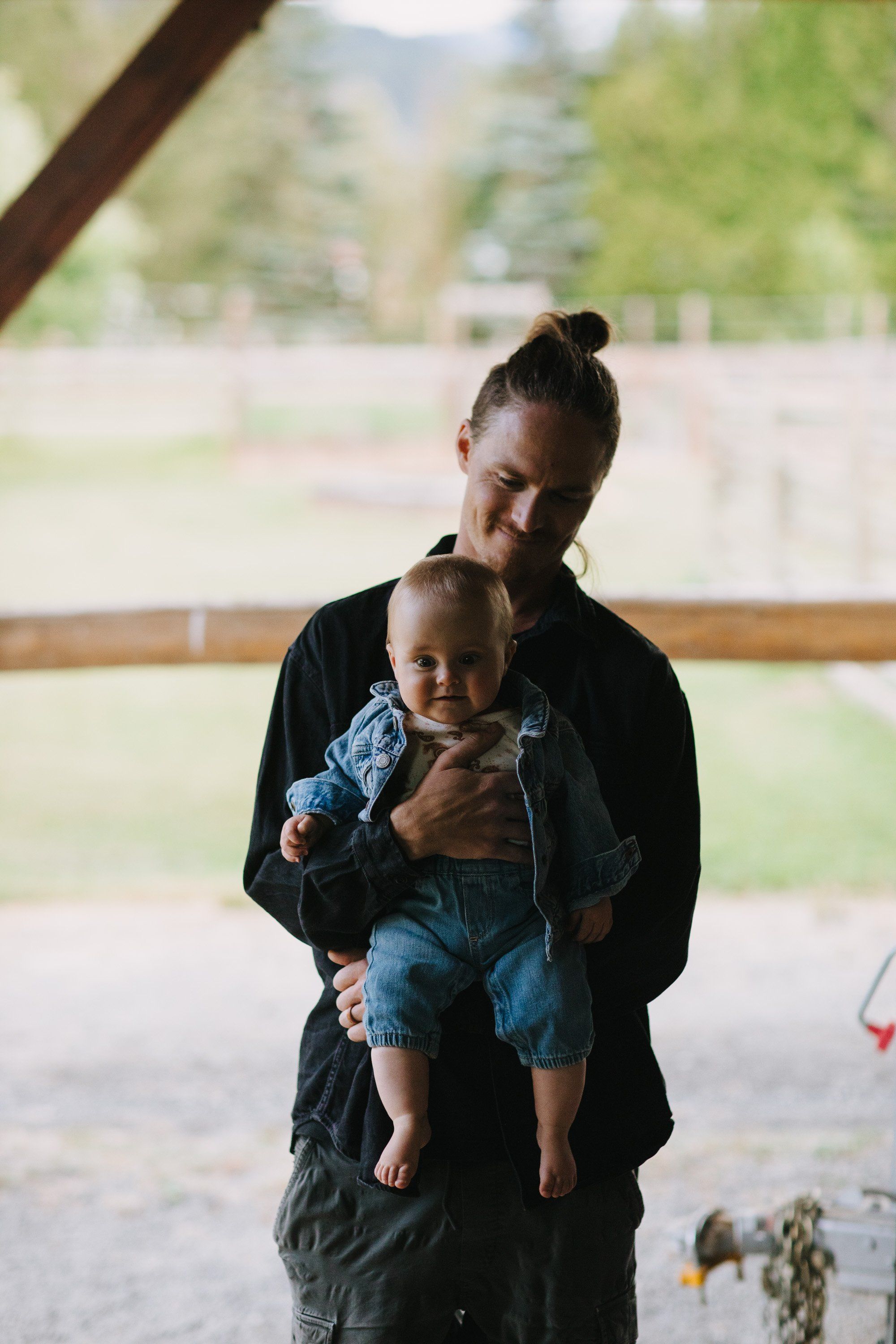 Mother holding young child in denim under a covered structure with forested mountains softly blurred behind them
