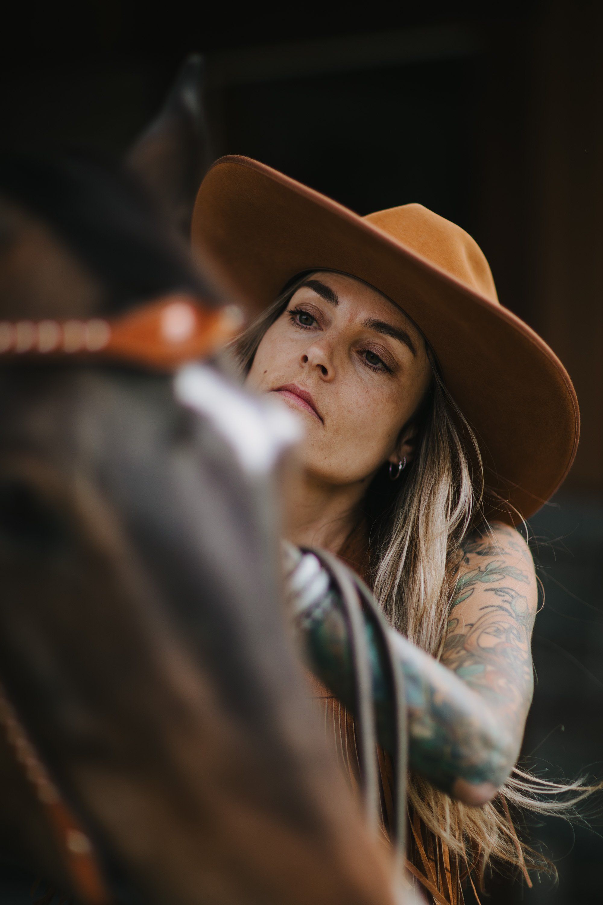Woman in brown cowboy hat with long hair and arm tattoos gazes upward beside a blurred horse, warm golden light against dark background