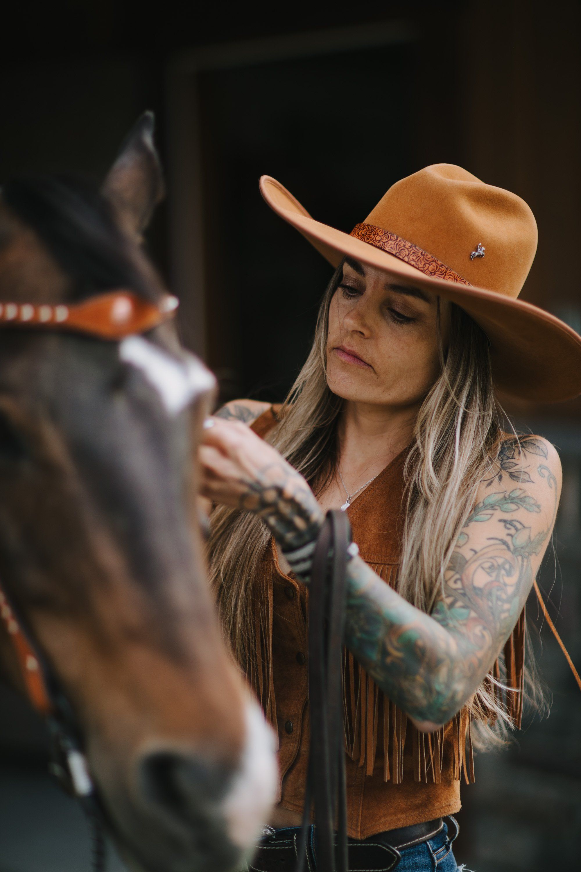 Woman in tan cowboy hat and fringe jacket holds horse's bridle against dark background, warm natural light on her face and tattoo