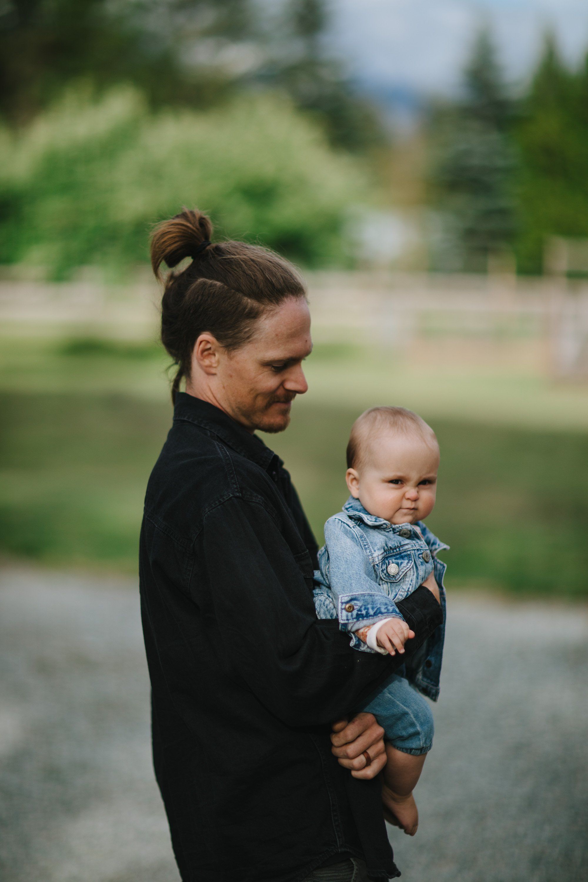 Woman holding toddler in denim jacket, both gazing toward forested mountains beside a calm lake