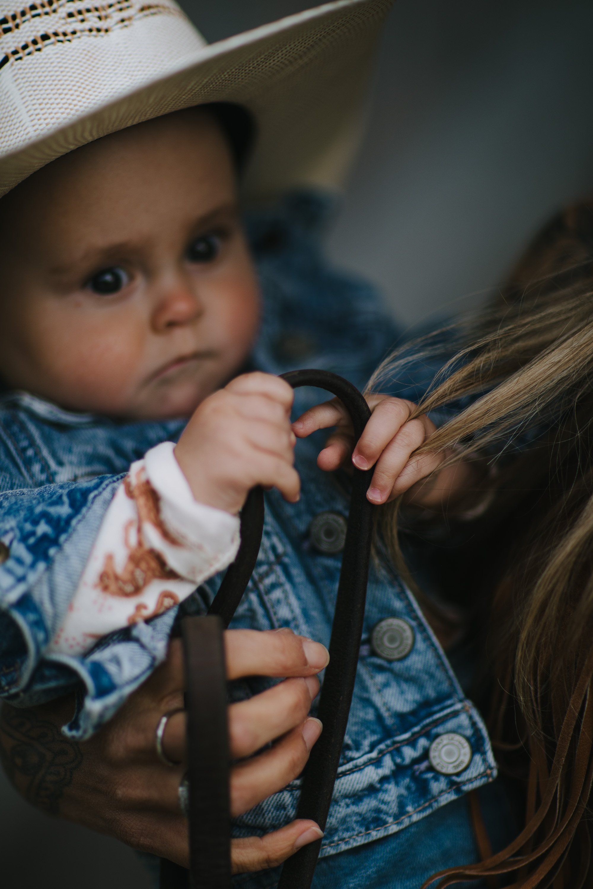 Toddler in cowboy hat and denim jacket grasps woman's hair with both hands, soft natural light against neutral background