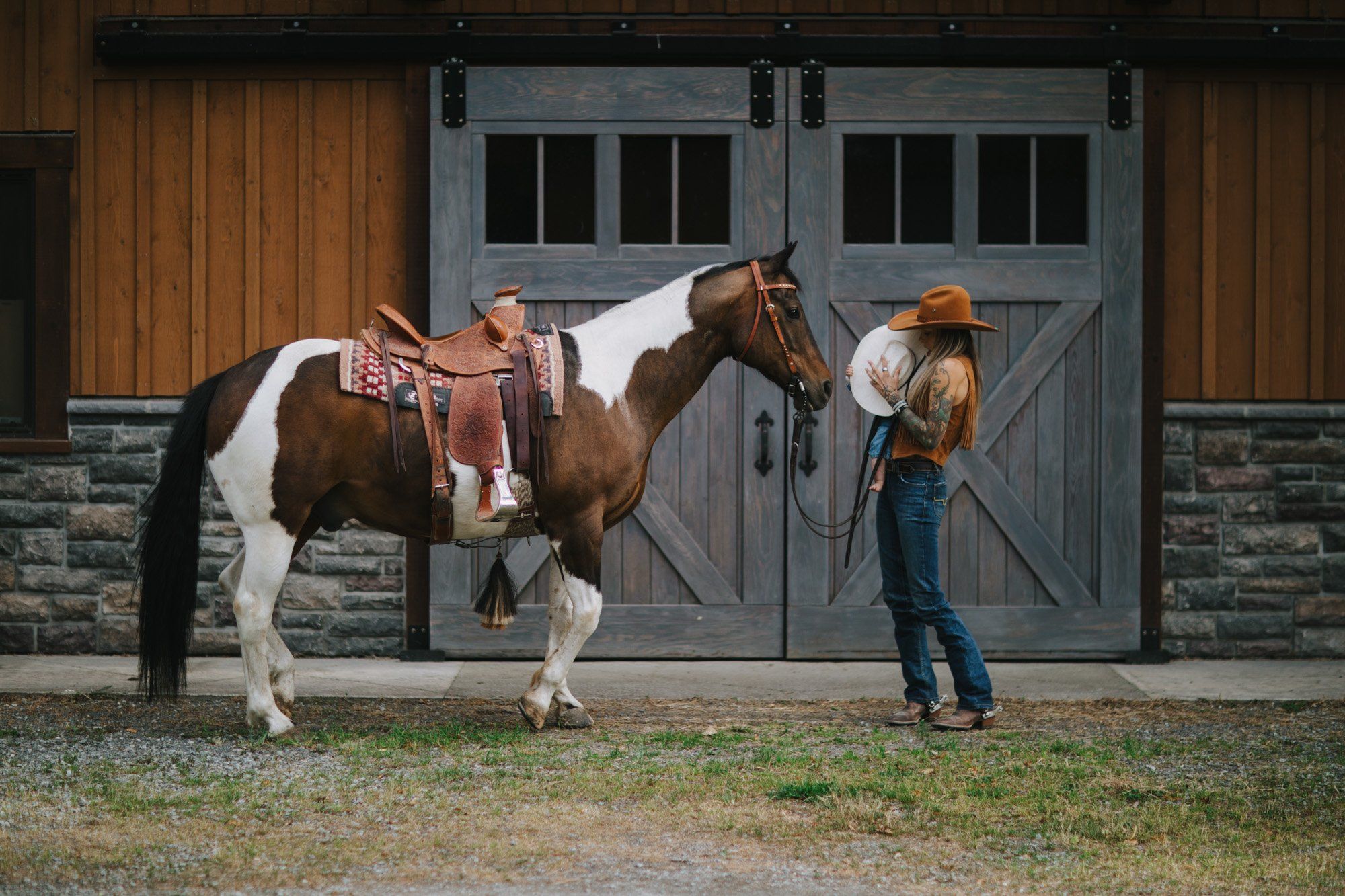 Cowboy in hat adjusts saddle on brown and white horse while woman in jeans holds reins beside rustic barn doors