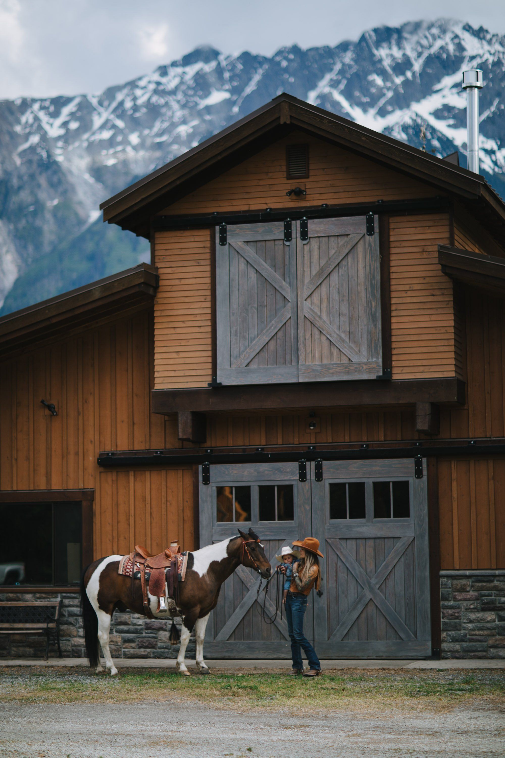 Woman in denim standing beside a saddled horse in front of a rustic barn with snow-capped mountains behind