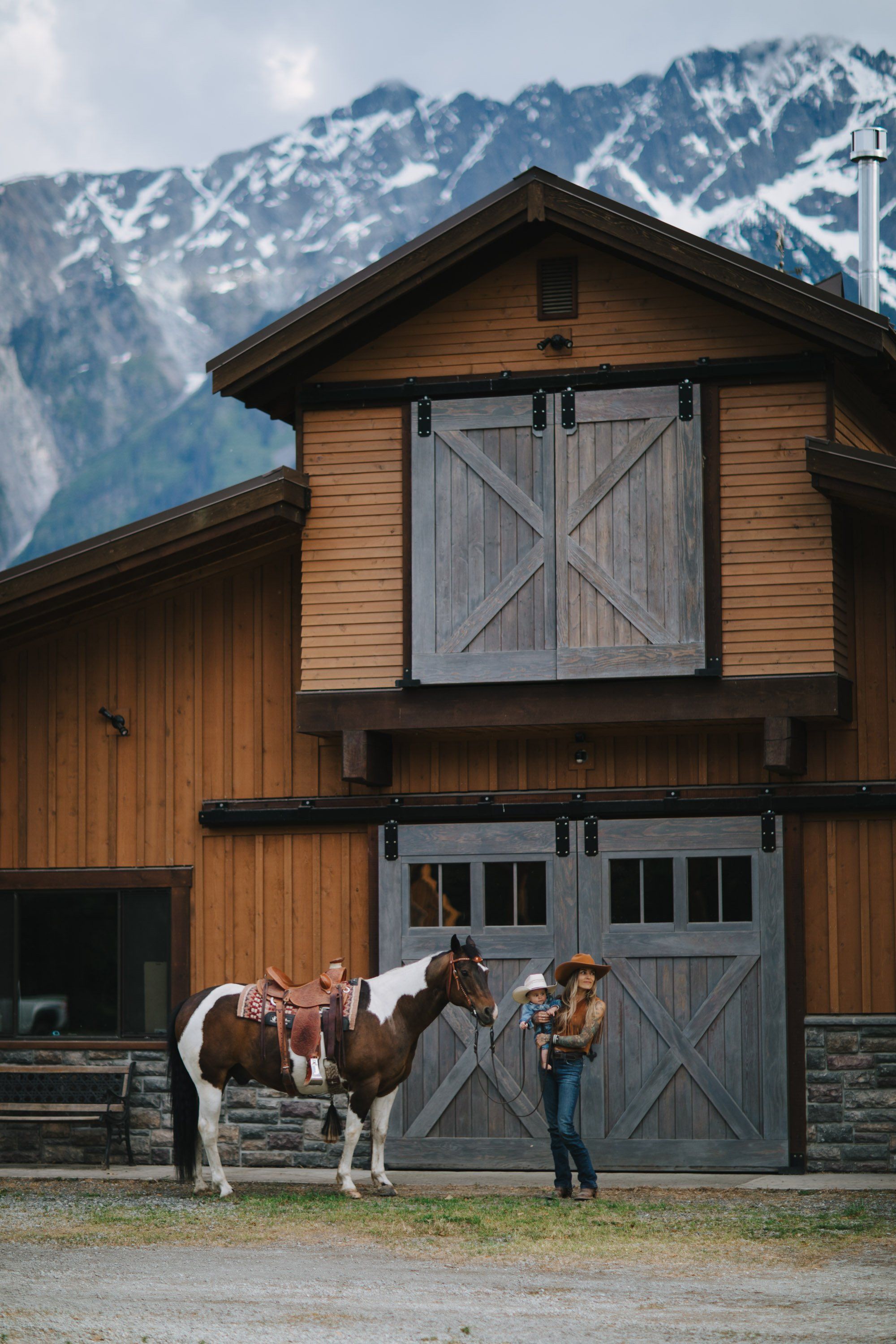 Woman standing beside a saddled brown horse in front of a timber barn with snow-capped mountains in the background