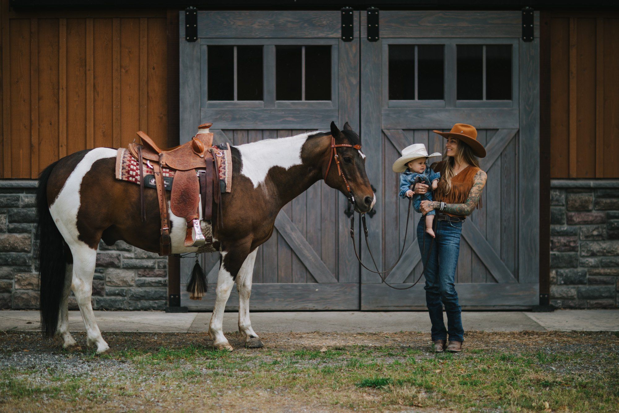 Woman in cowboy hat holds child while standing beside saddled brown and white horse in front of rustic barn doors