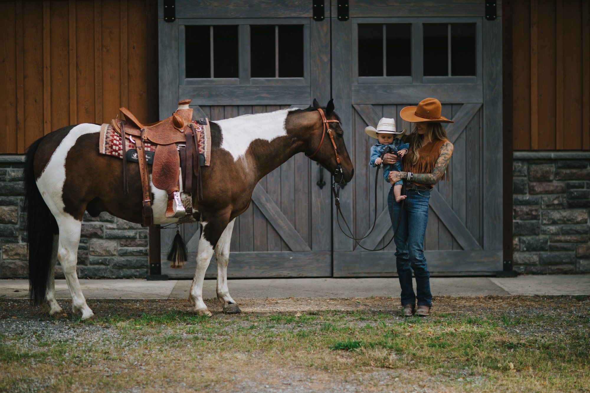 Cowgirl holds horse's bridle while posing in front of rustic barn doors in golden afternoon light
