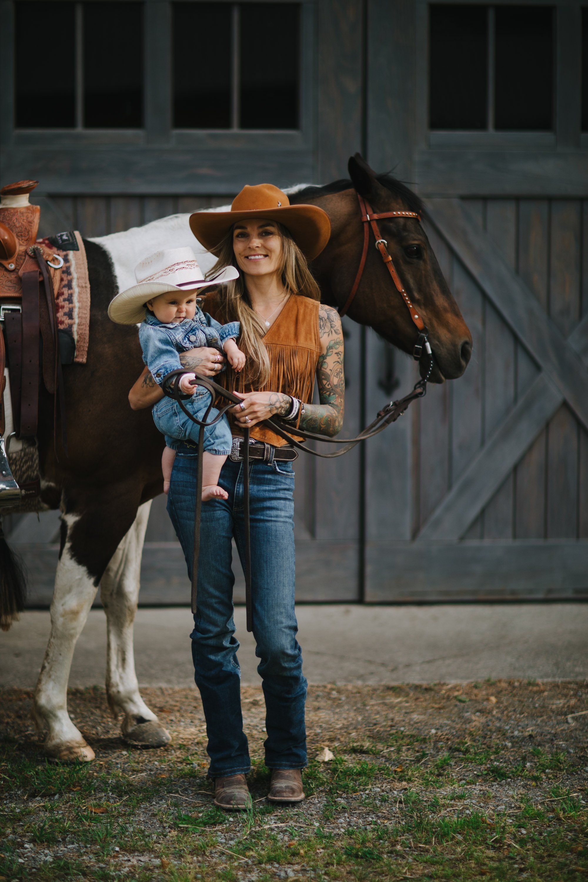 Woman in cowboy hat holding toddler beside saddled horse and man in barn setting, golden hour light