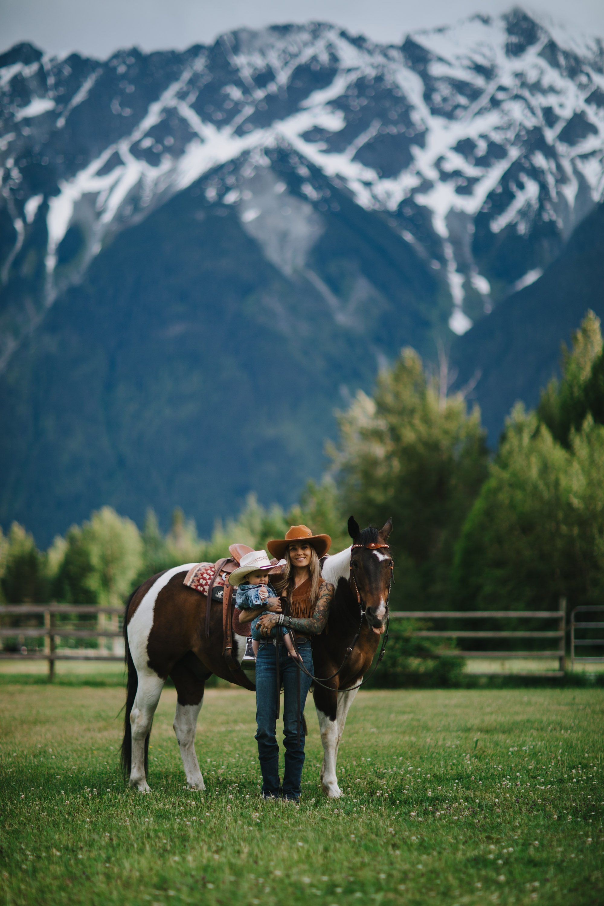Cowgirl stands with saddled brown and white horse in alpine meadow, snow-capped peaks rising behind evergreen forest
