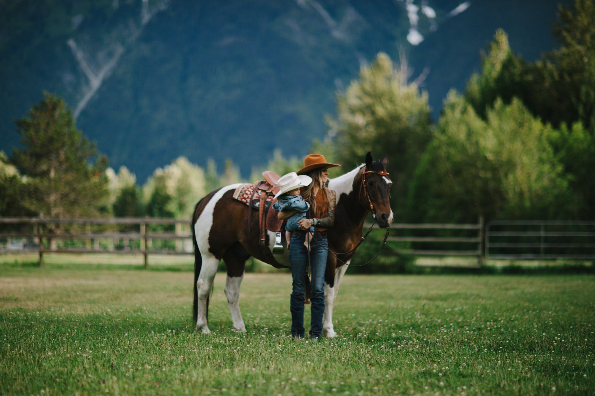 Cowgirl in denim stands beside a brown and white horse in a sunlit meadow with evergreen trees and mountains under blue sky