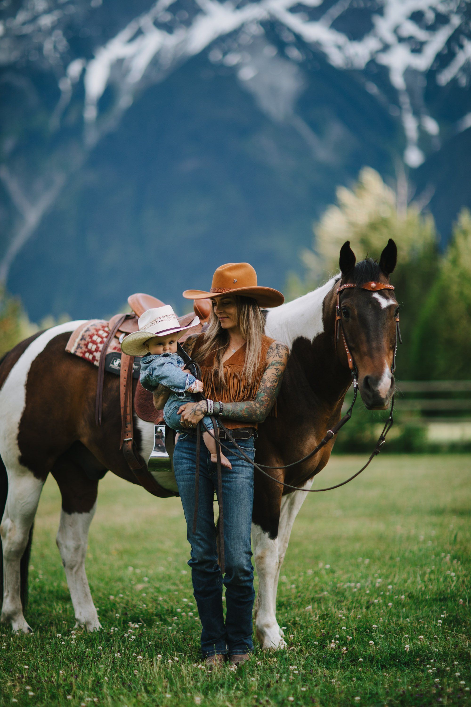 Woman in cowboy hat and denim stands with saddled horse against snow-capped mountain backdrop