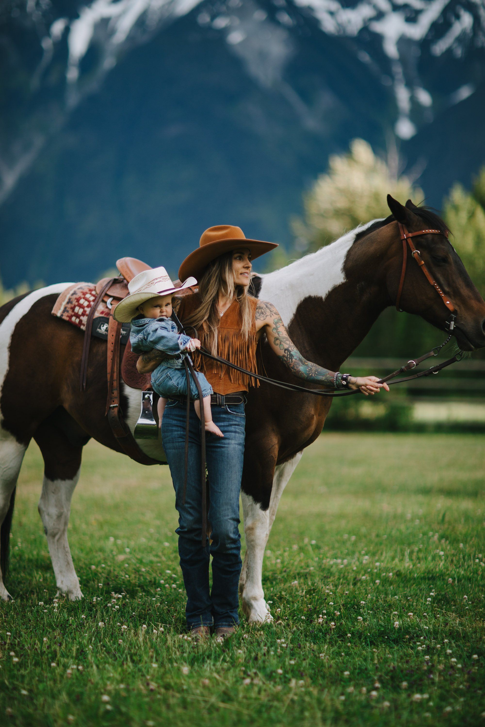 Young child in cowboy hat and denim sits on saddled horse with mountains and green meadow behind