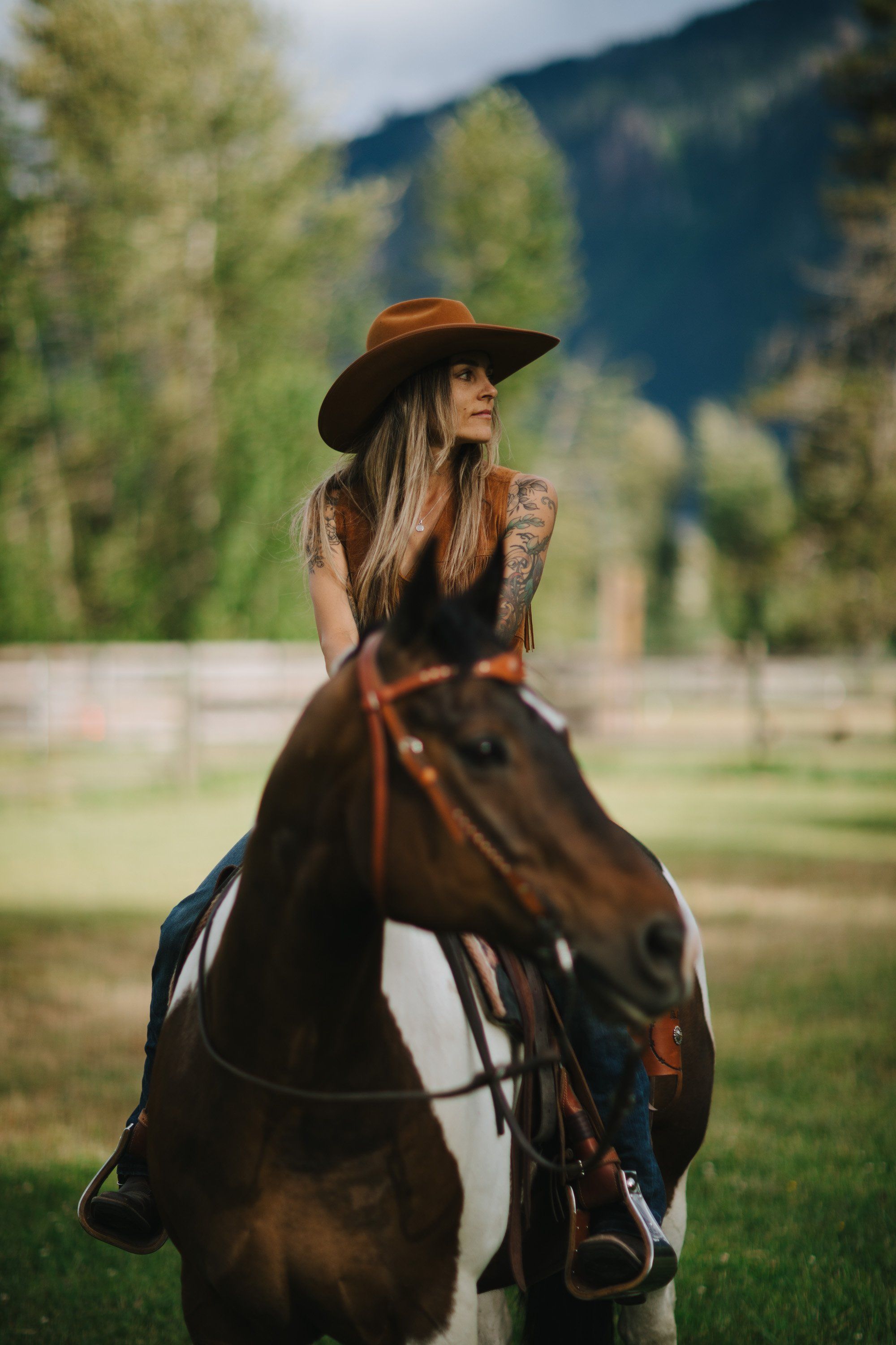Woman in rust cowboy hat sits sidesaddle on brown horse in sunlit meadow with evergreen forest and mountains behind