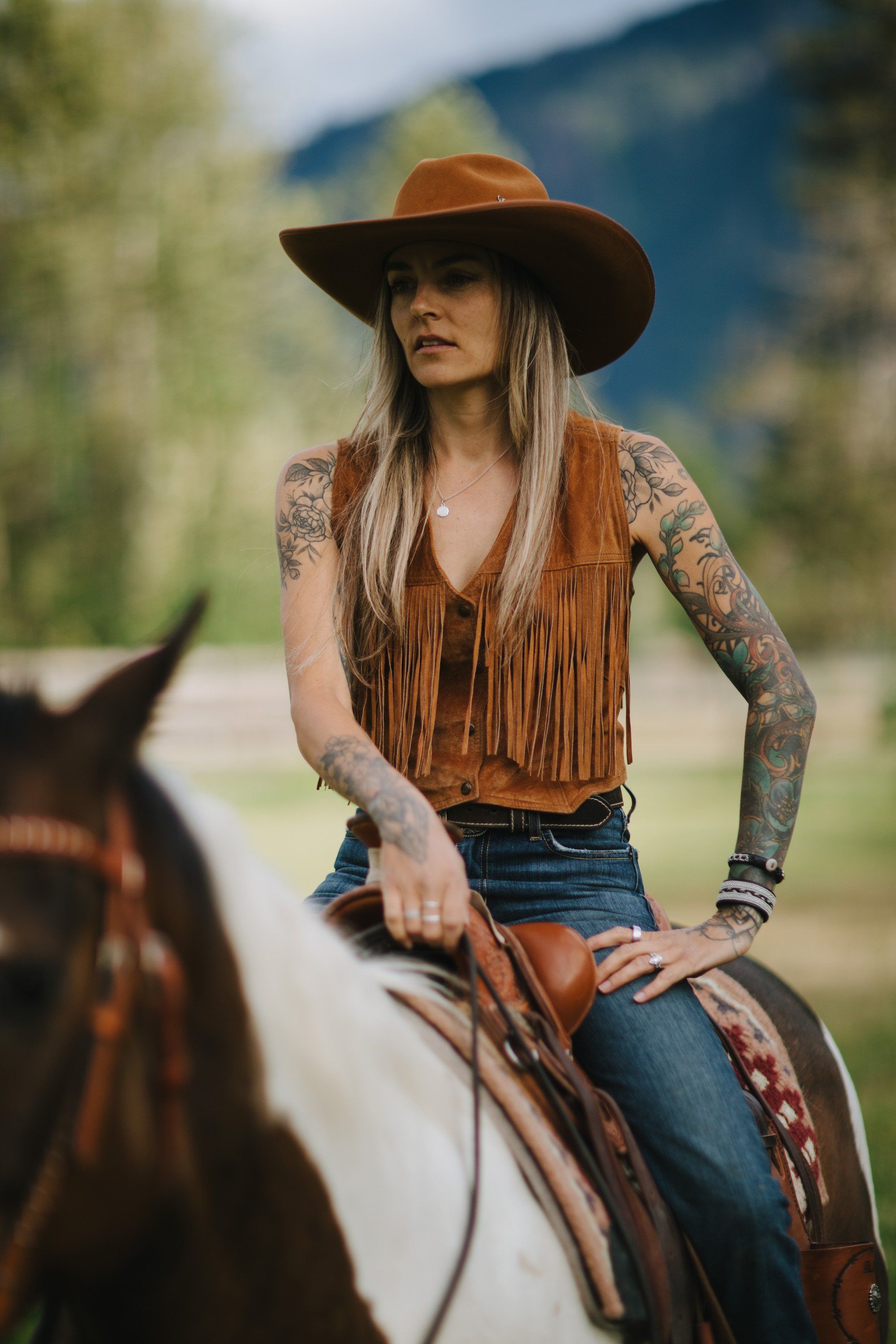 Woman in fringe suede vest and cowboy hat sits on horseback in mountain valley, long hair flowing, afternoon light