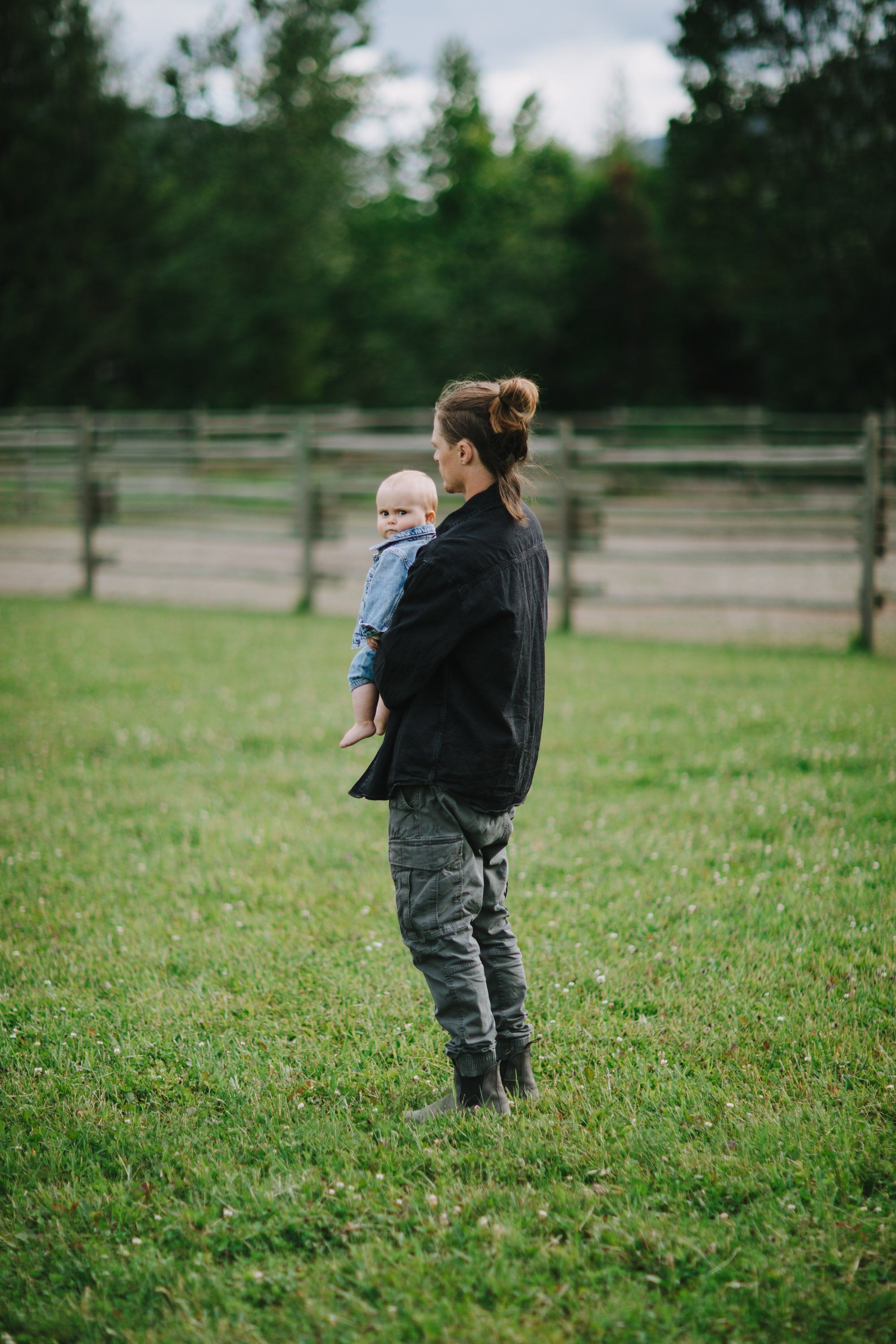 Woman holding infant standing in grassy field with wooden fence and evergreen trees under overcast sky