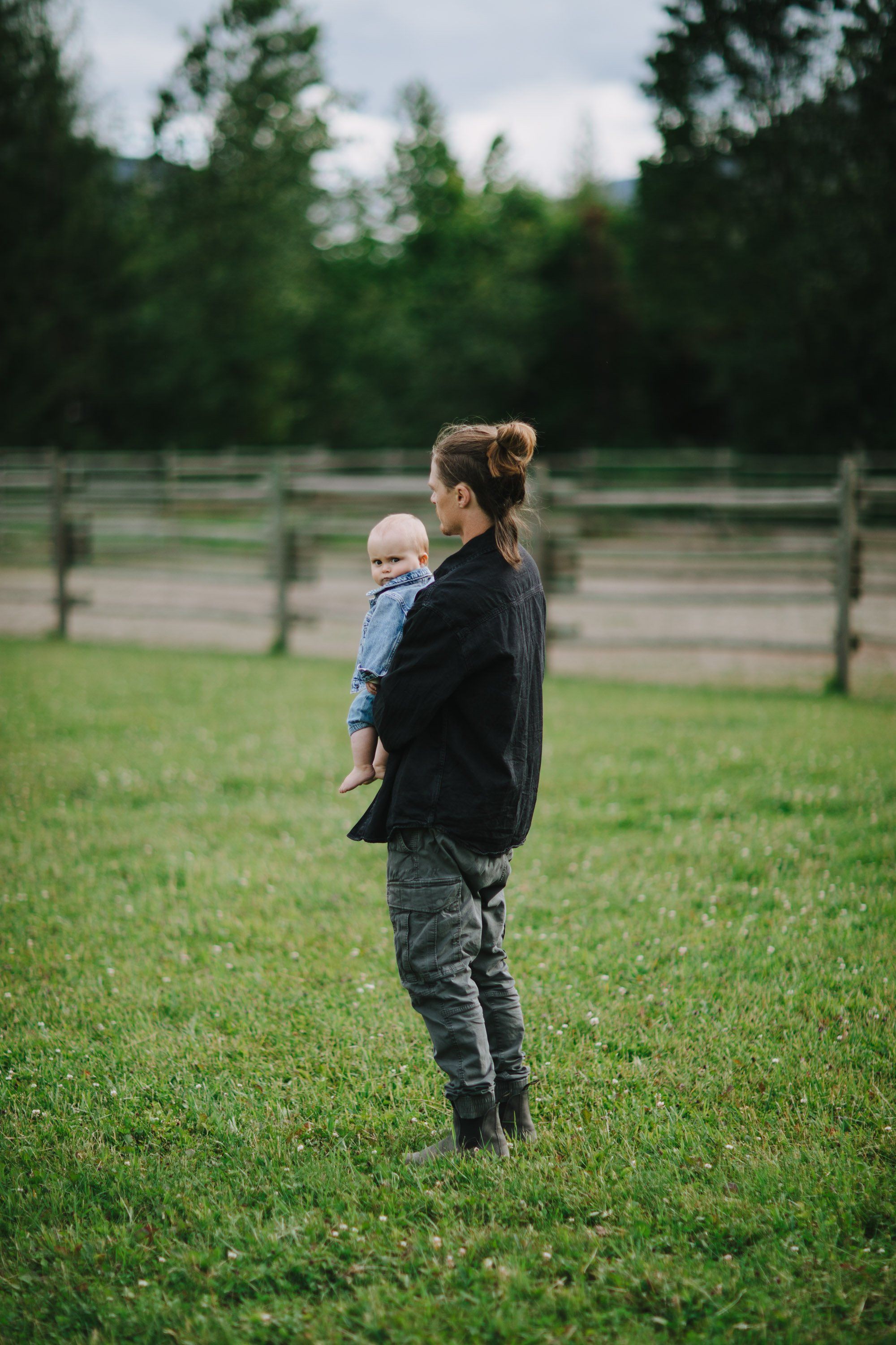 Woman holding toddler standing in a grassy field with wooden fences and evergreen trees under soft overcast light