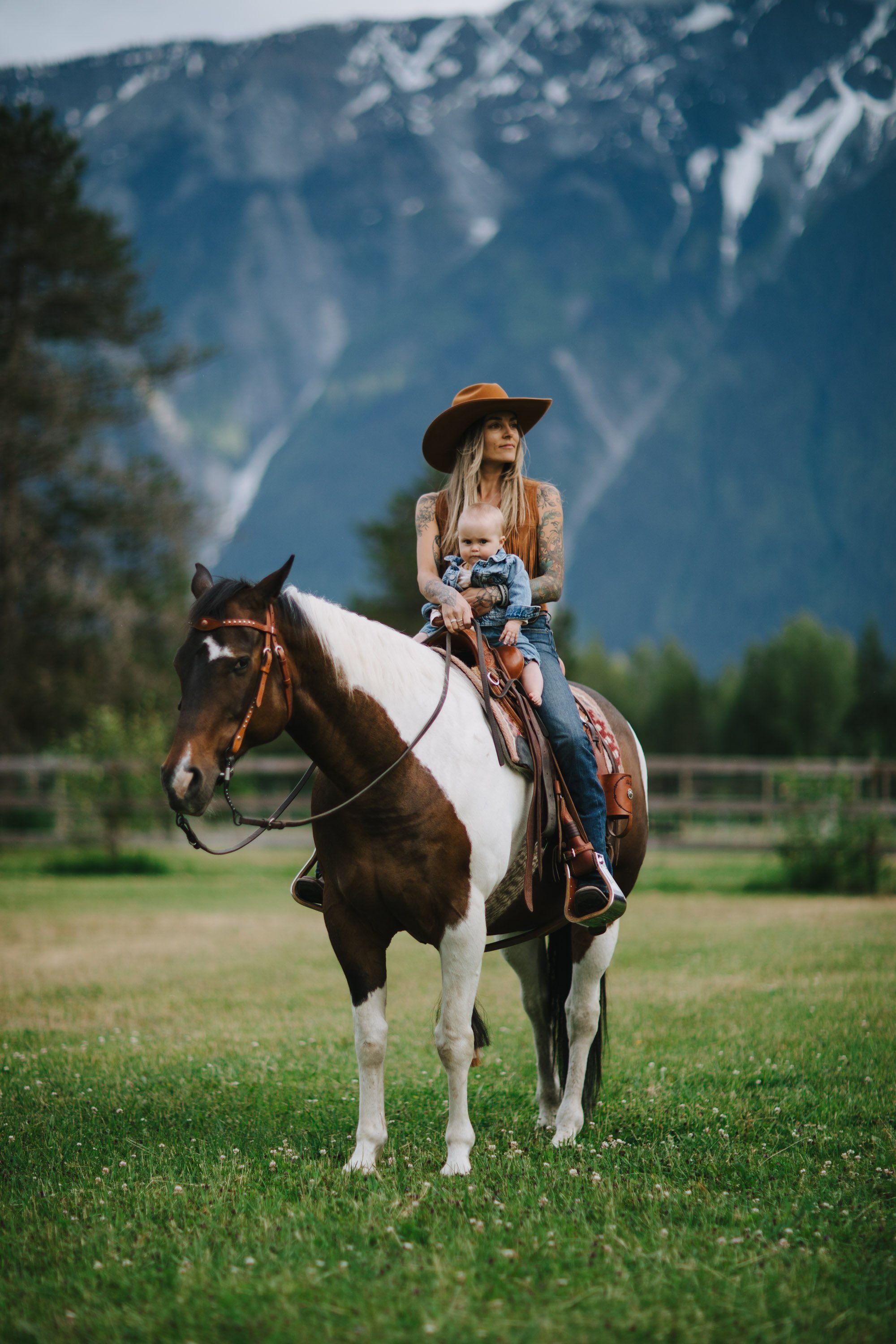 Woman in cowboy hat on brown and white horse with mountain backdrop in grassy field