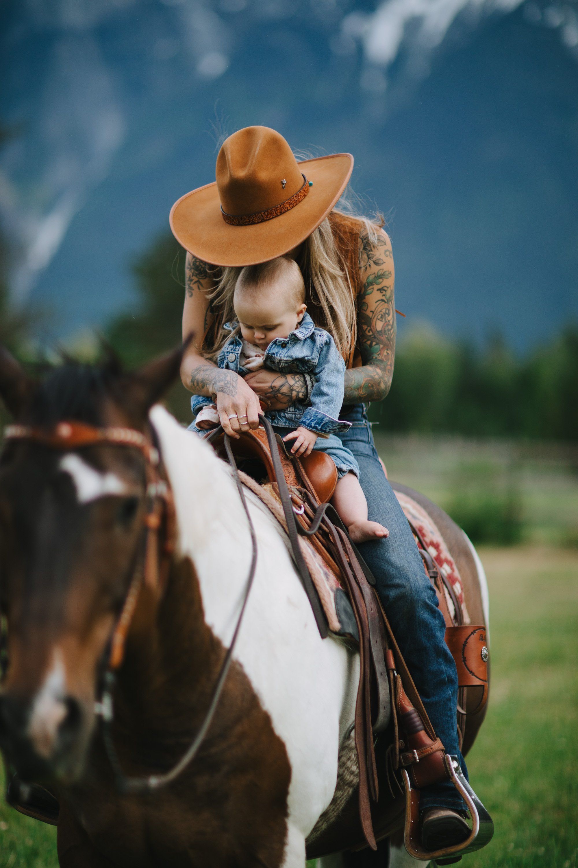 Woman in cowboy hat and denim riding a brown-and-white horse against blue sky and mountain backdrop
