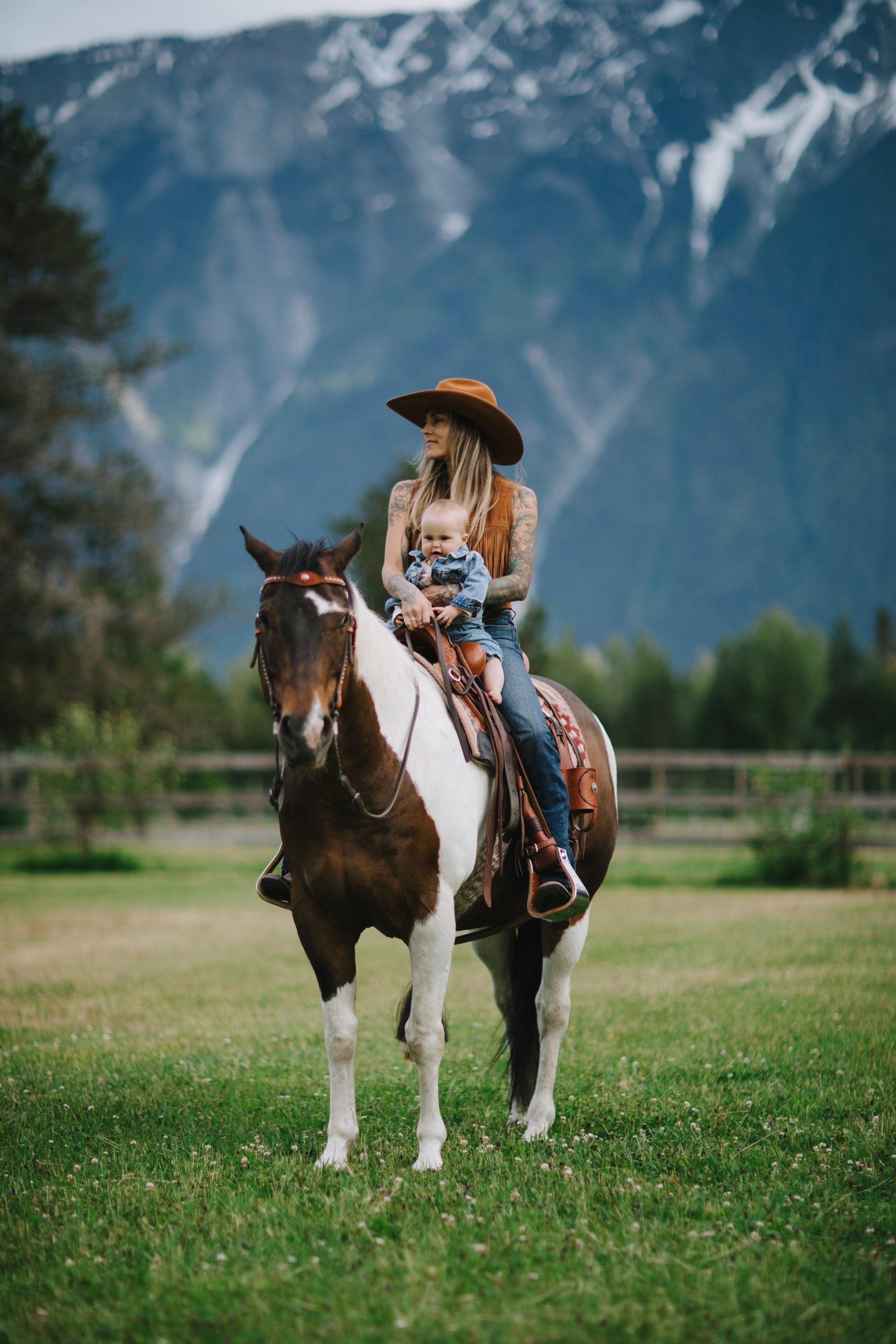 Woman in cowboy hat on brown and white horse with mountains and green meadow in background