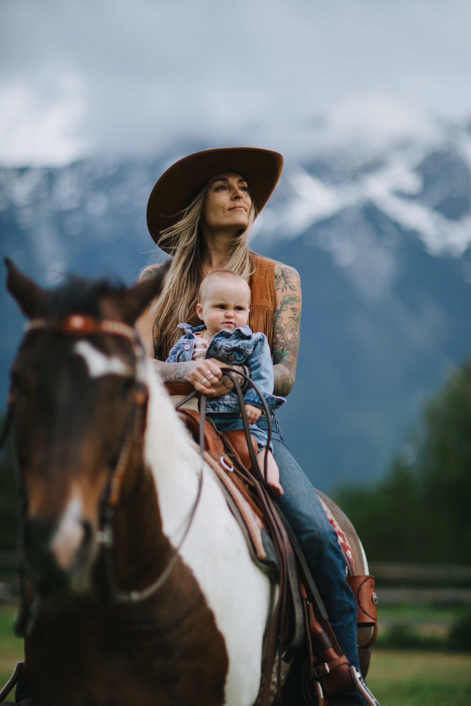 Woman in cowboy hat holding infant on horseback with snow-capped mountains and evergreen forest in soft, overcast light