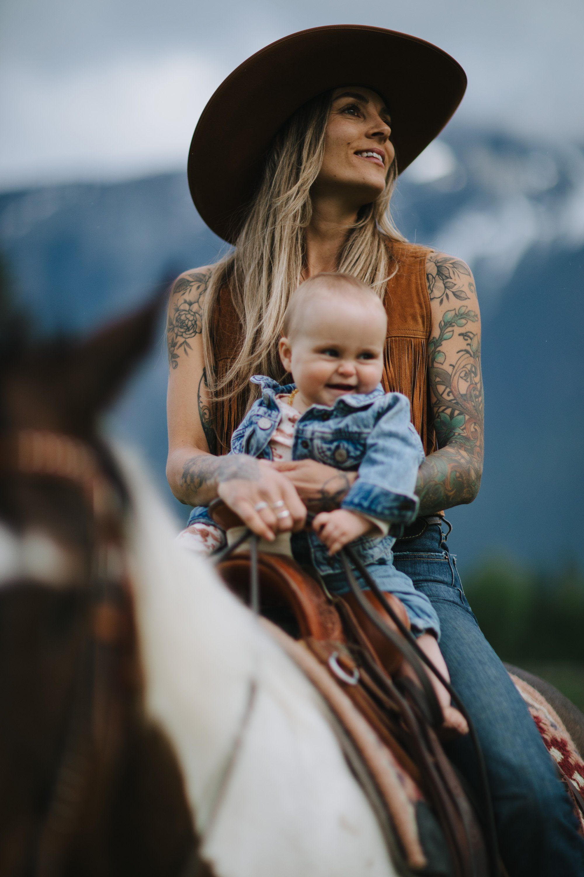 Woman in black cowboy hat holding smiling baby on horseback with misty mountains behind