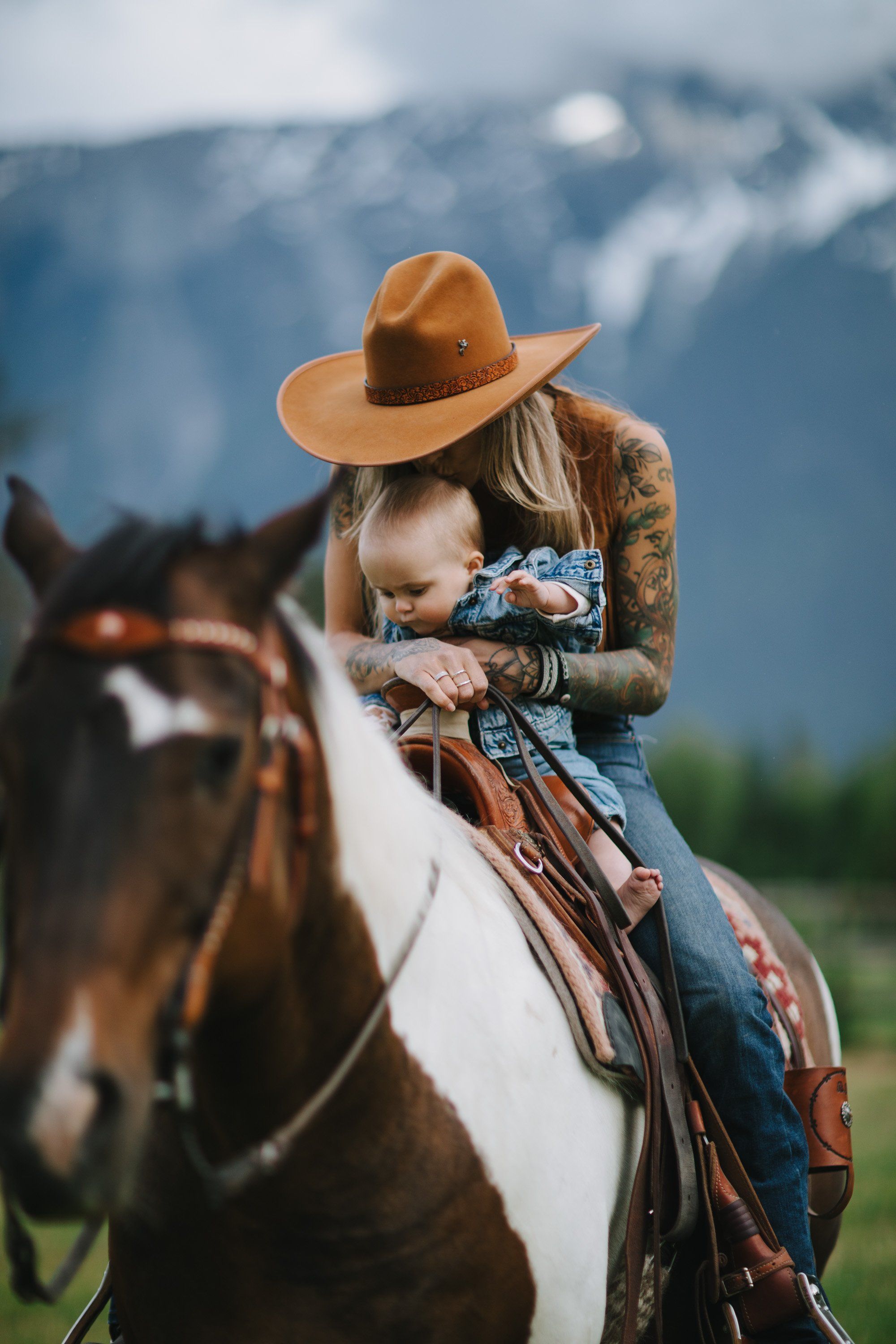 Woman in tan cowboy hat and denim on horseback with mountains in soft focus behind her