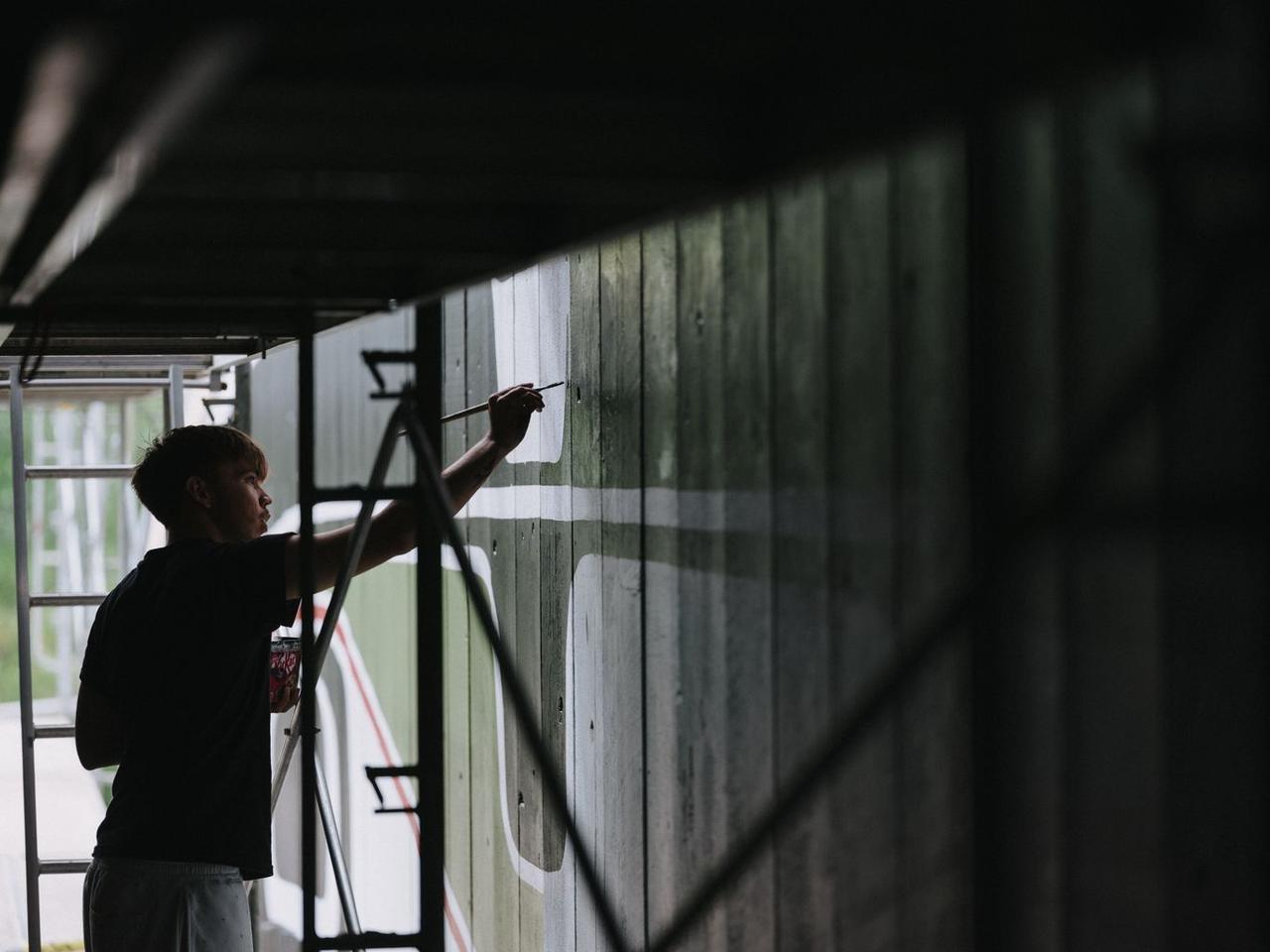 Hand holding paintbrush poised at weathered wooden wall panels during repainting of Namgis First Nation Big House