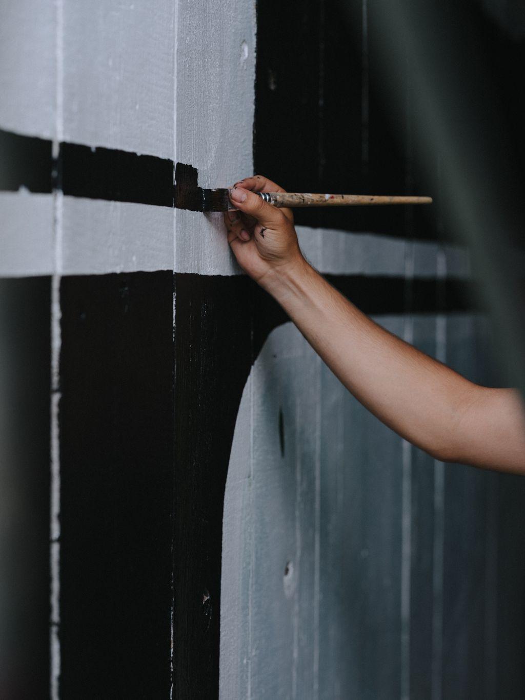 Hand with brush applying black paint to light gray wall in geometric pattern