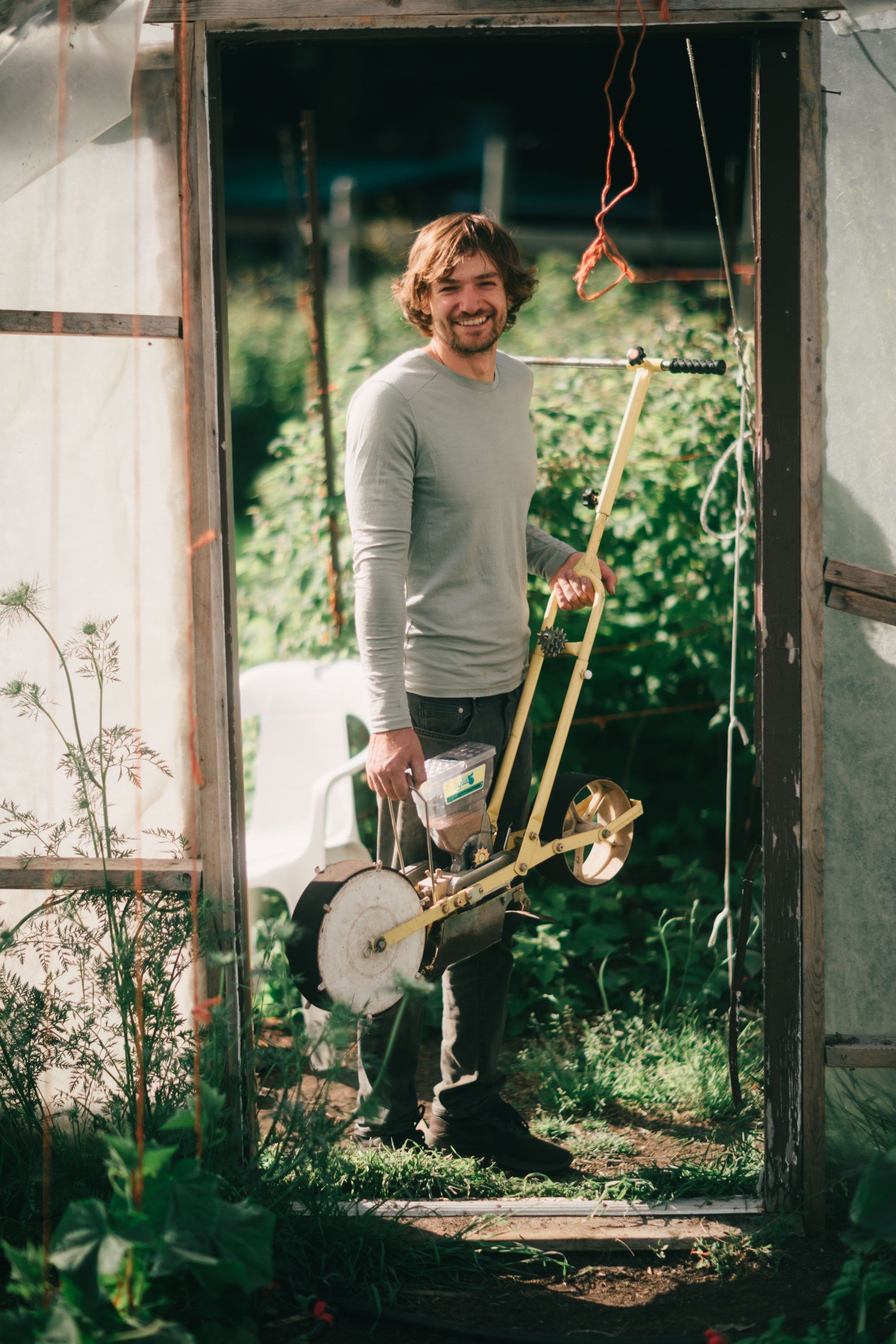 Man smiling in greenhouse doorway holding farming tool, lush garden visible behind him in dappled sunlight
