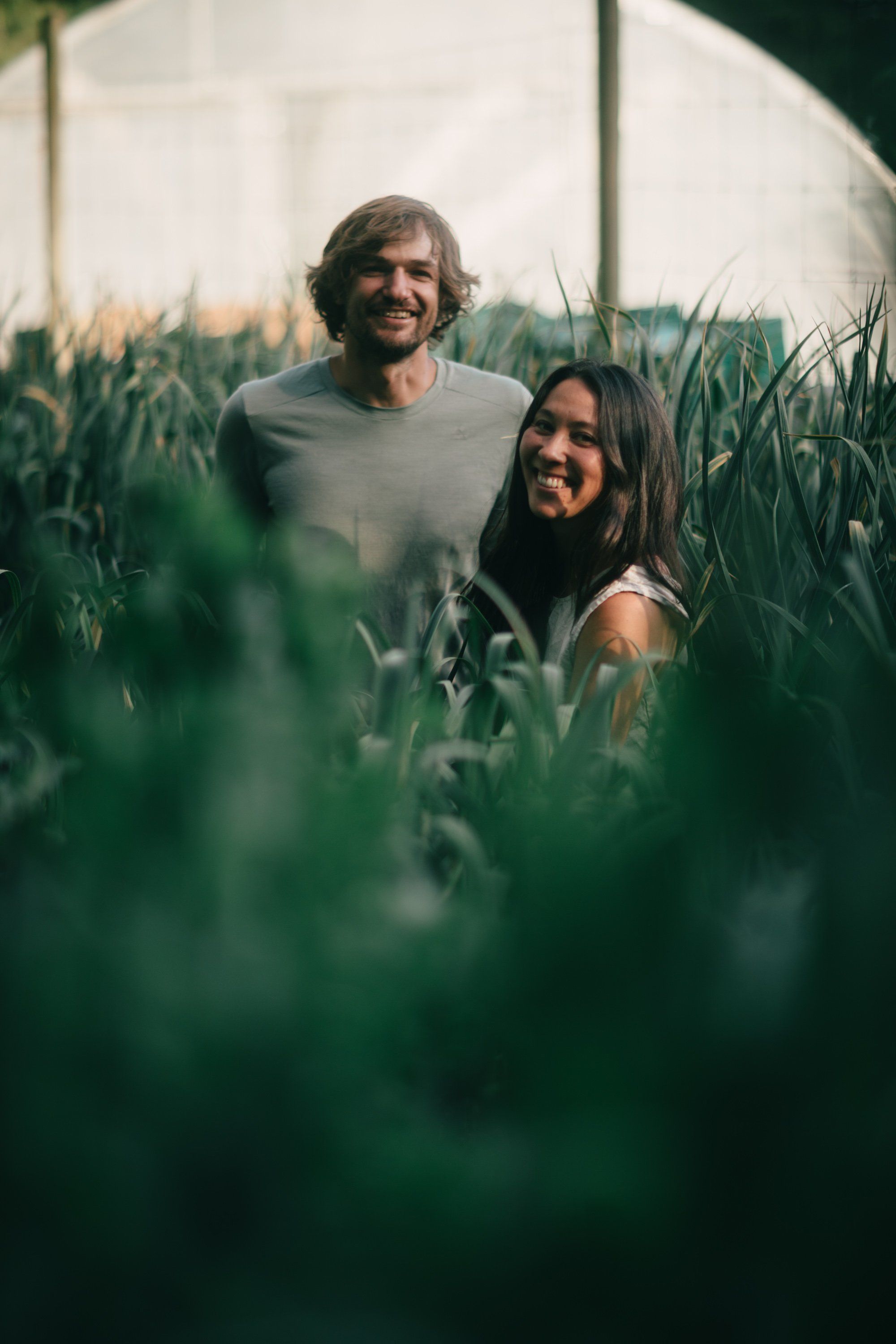 Couple smiling among tall green plants in a sun-dappled greenhouse with soft natural light filtering through