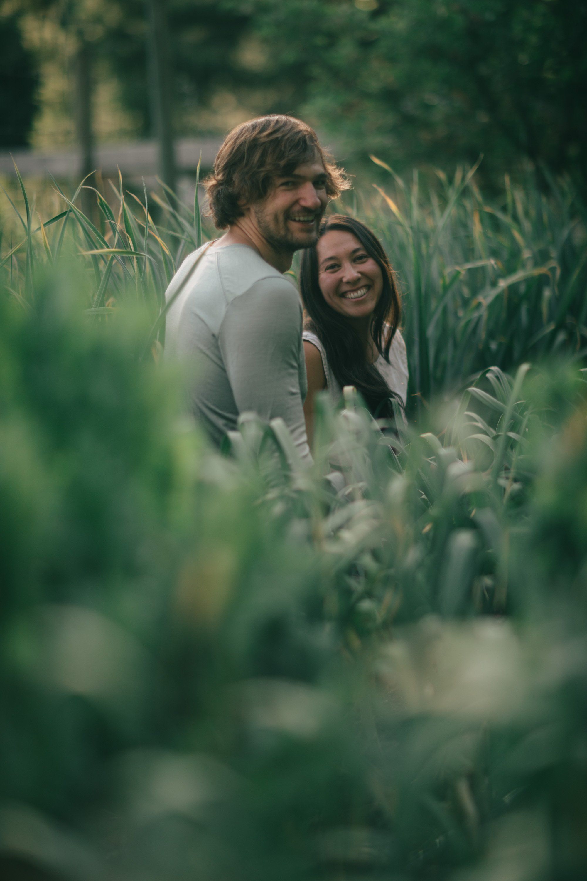 Couple nestled in tall green crops, smiling at each other in warm, dappled farmland light