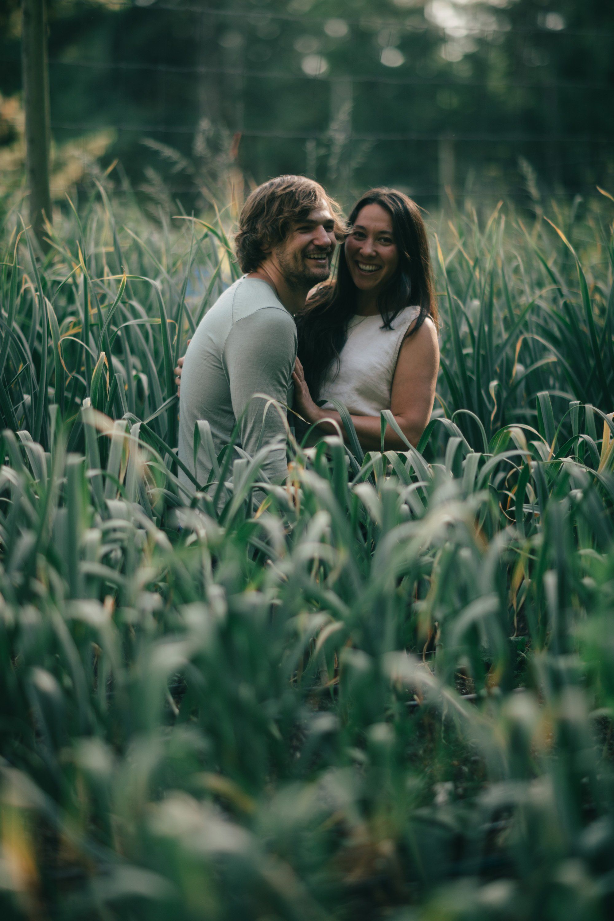 Couple laughing together among tall green crops in dappled sunlight, intimate moment in a rural farm setting