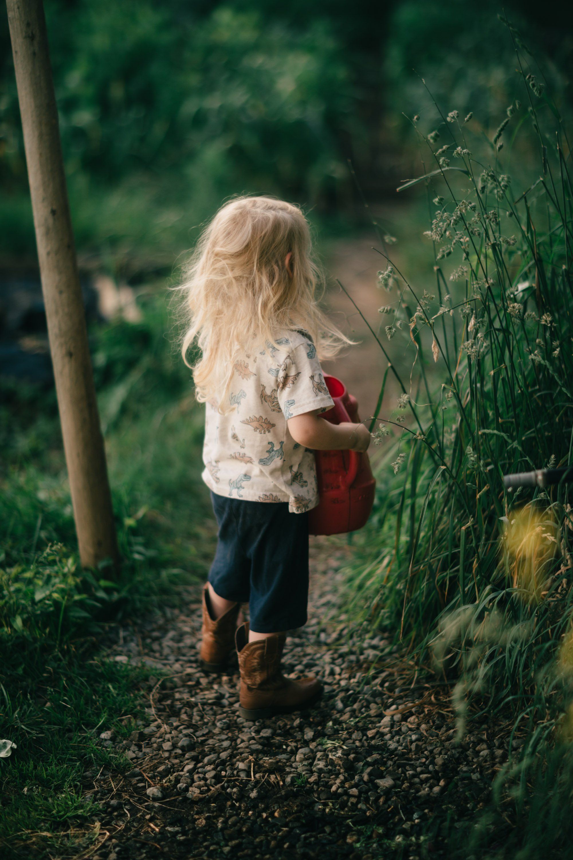 Child in white shirt with red backpack walking away down a leafy garden path dappled with natural light