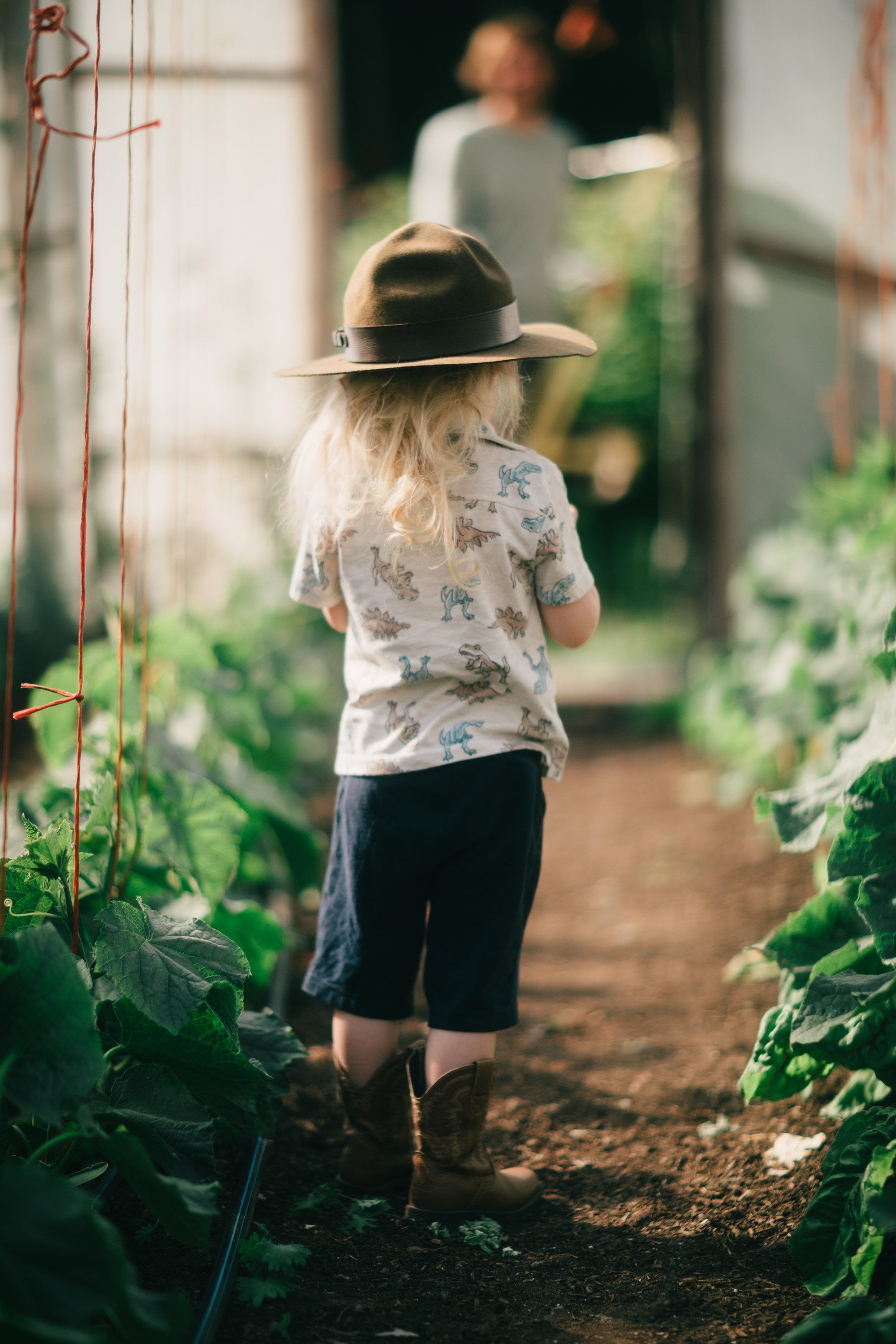 Child in floral shirt and hat walks barefoot down garden row lined with green plants and wooden stakes