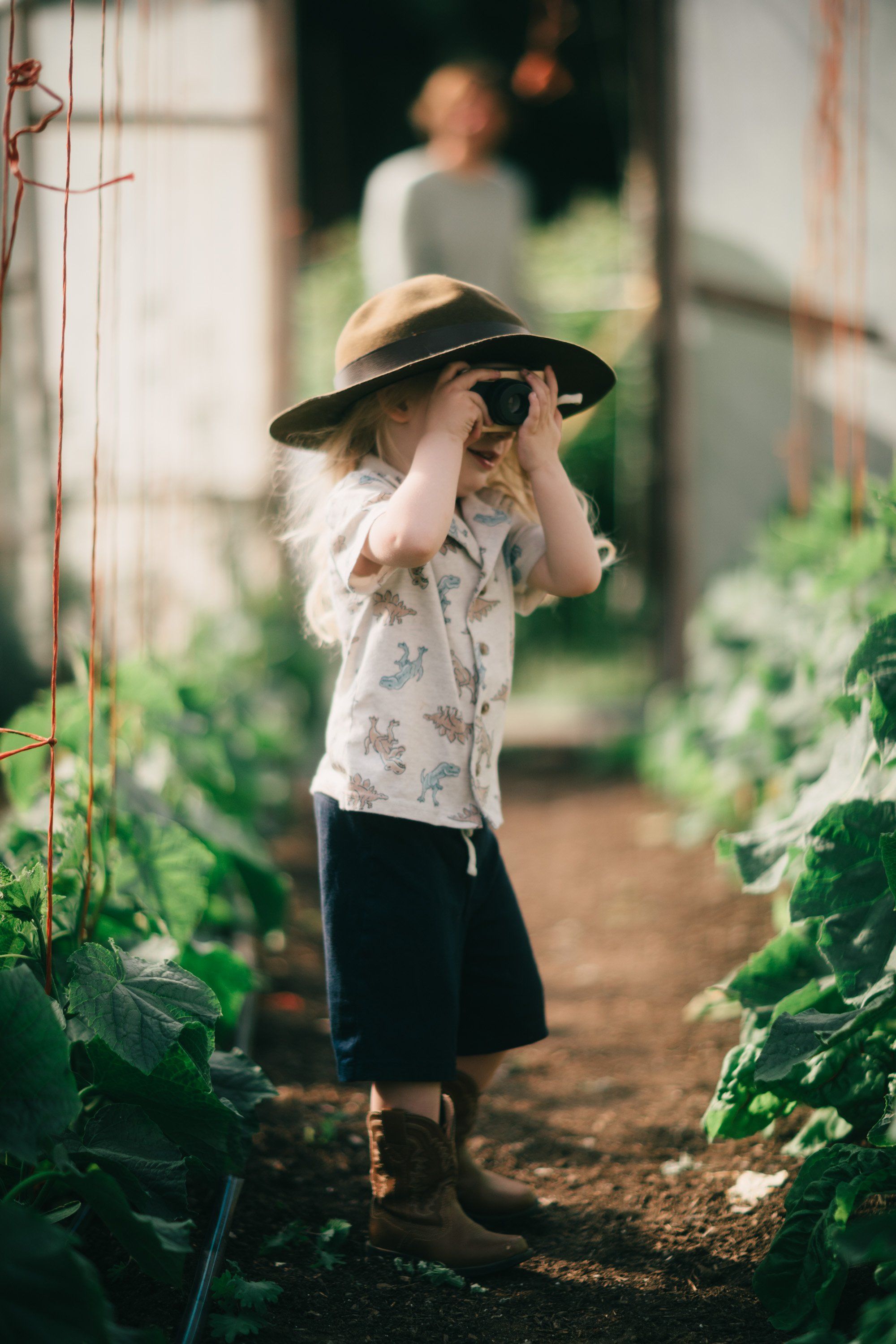 Child in floral shirt and straw hat adjusts sunglasses while standing between garden plants on a sun-dappled farm path