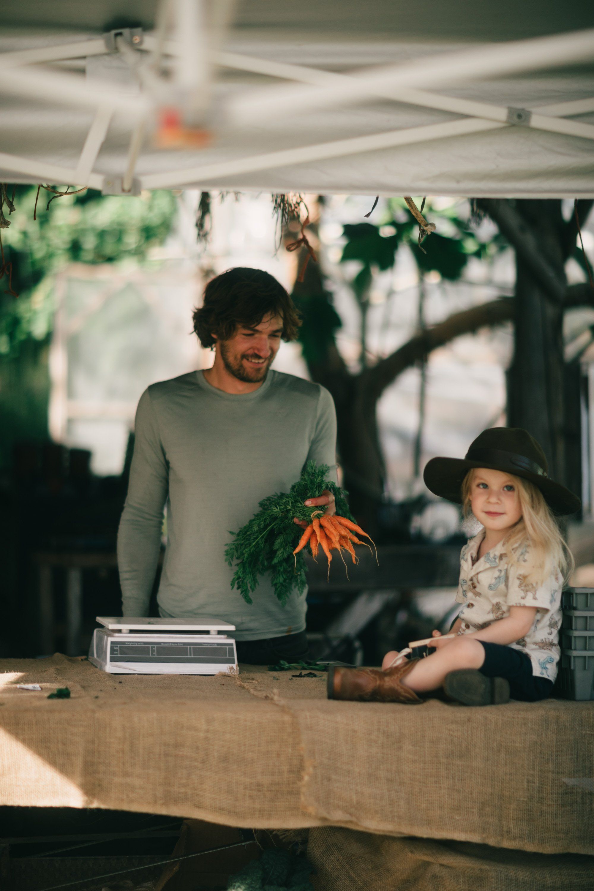 Man and child at farm stand holding fresh carrots under tent canopy with soft natural light