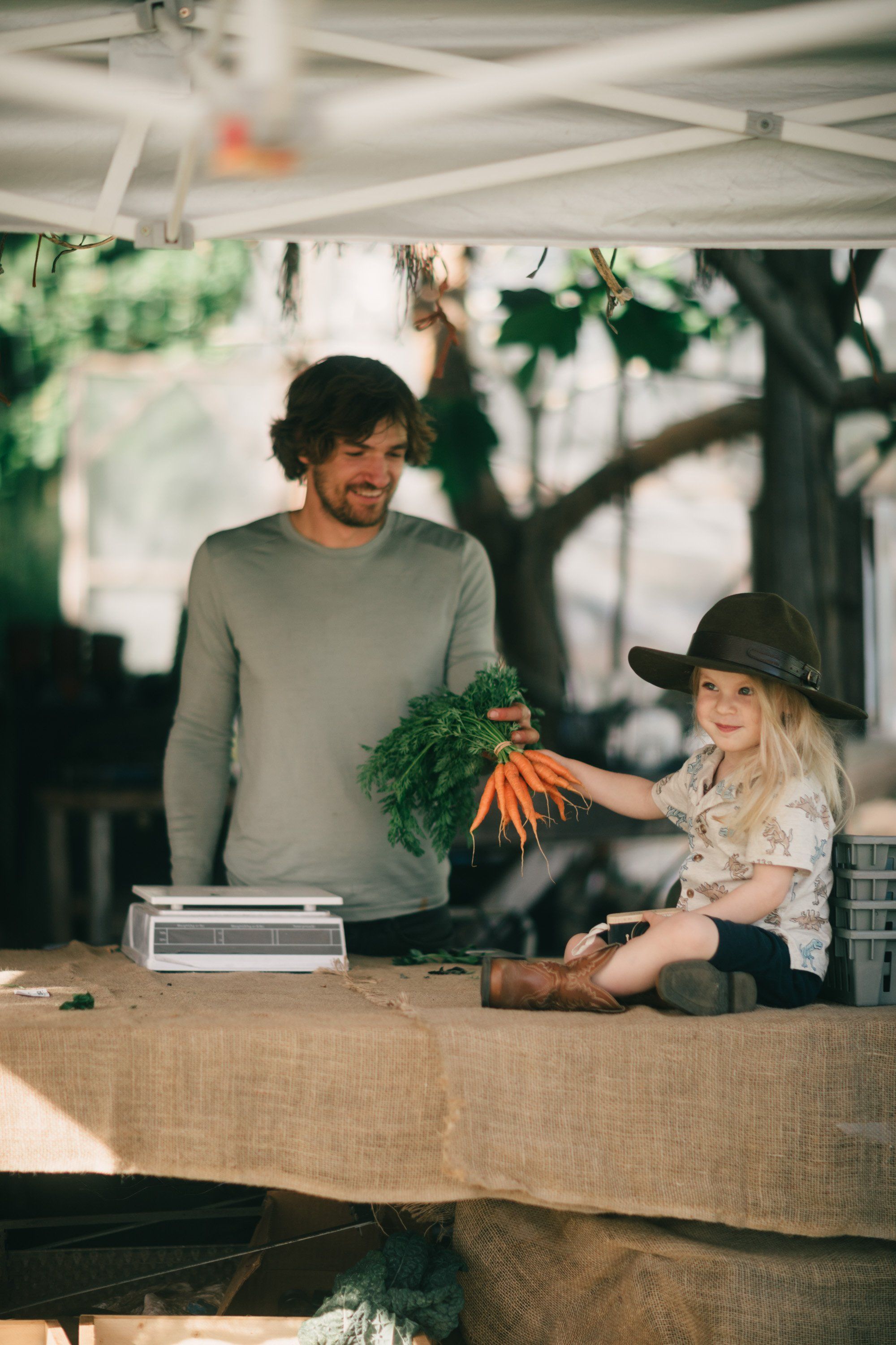 Man and child at a farm stand holding fresh carrots under dappled sunlight filtering through canvas canopy