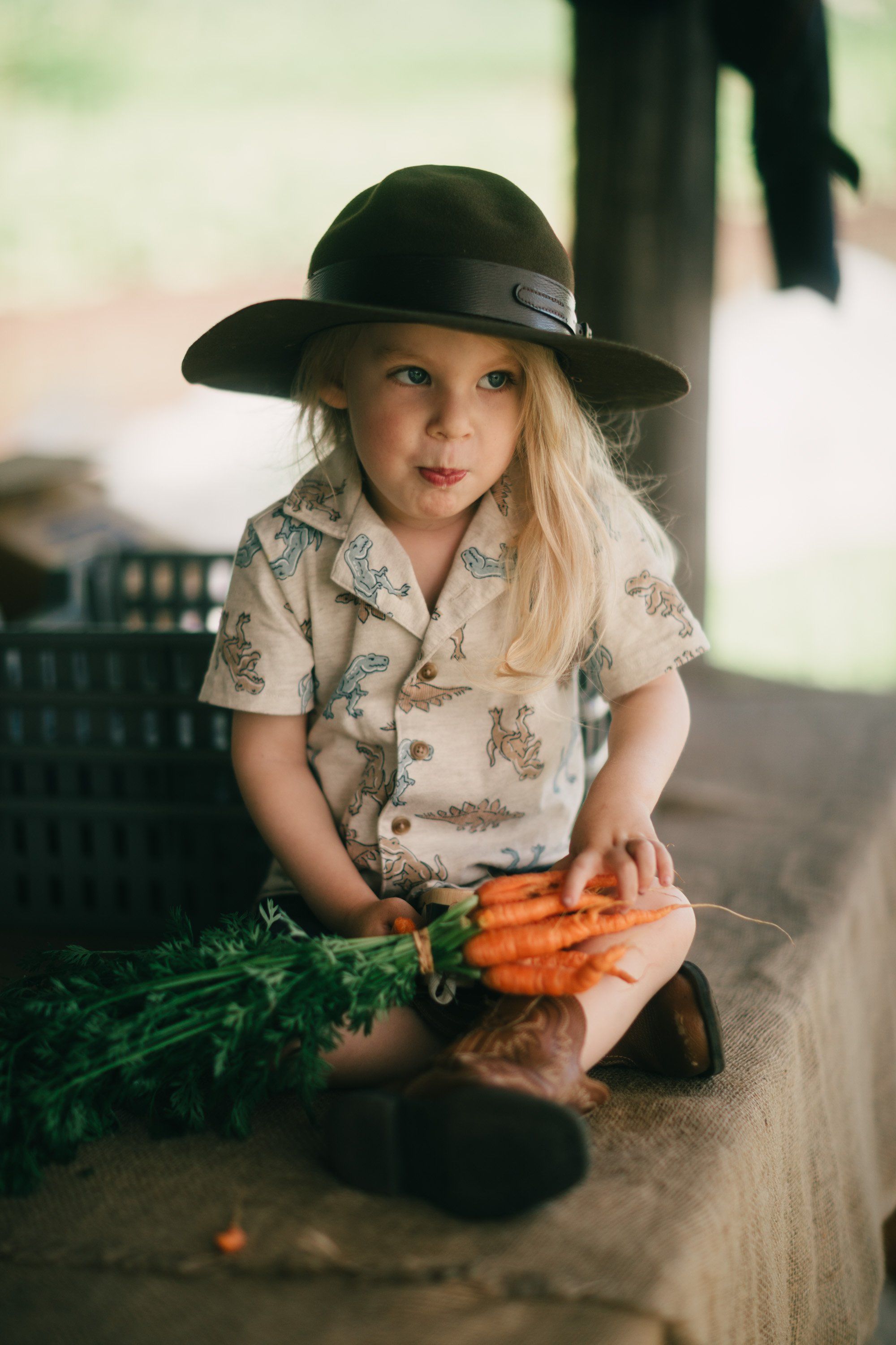 Young child in cowboy hat holding fresh carrots with green tops at a rustic farm setting