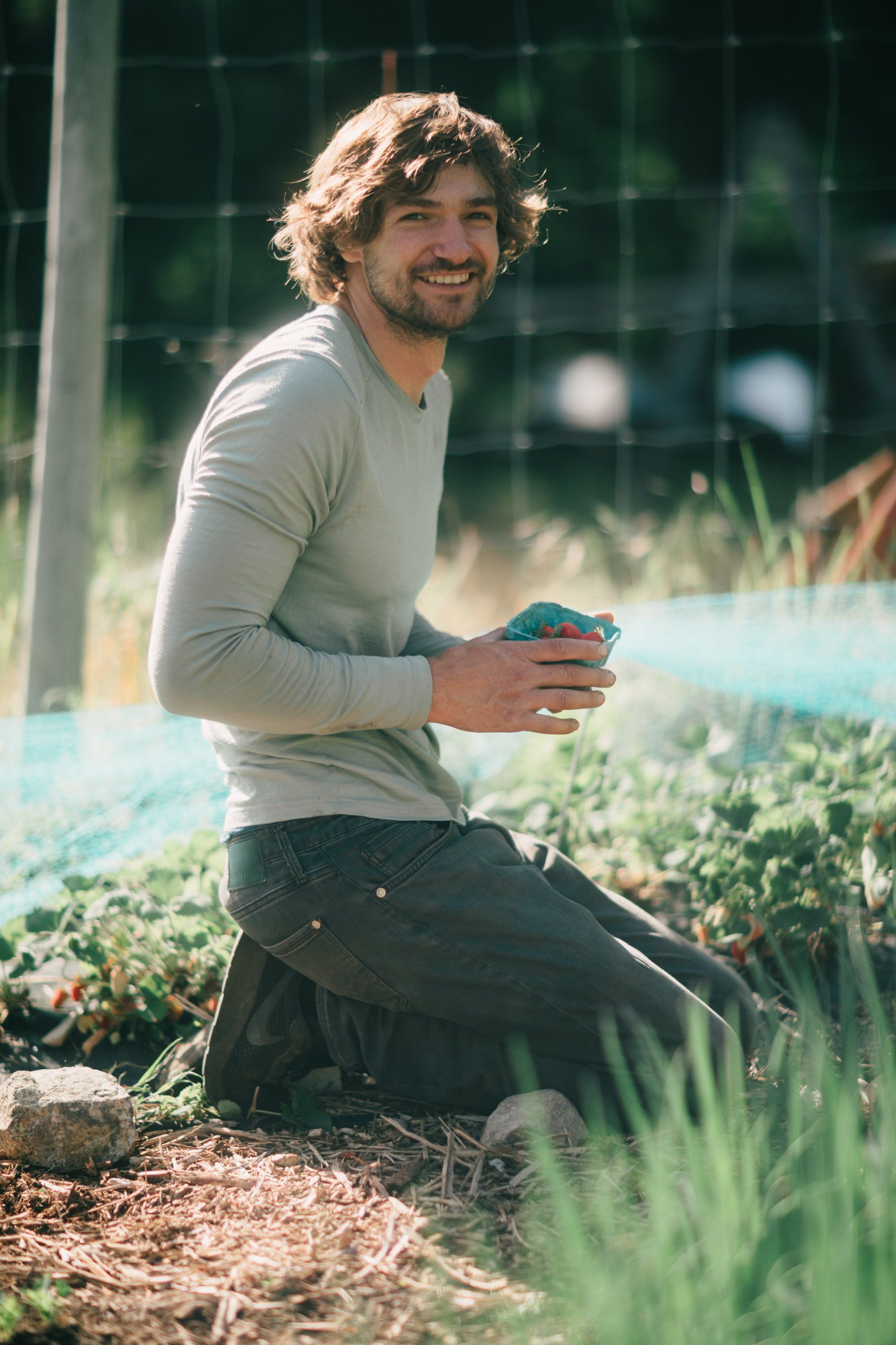 Man kneeling in garden holding seedling, surrounded by green plants and greenhouse, golden afternoon light