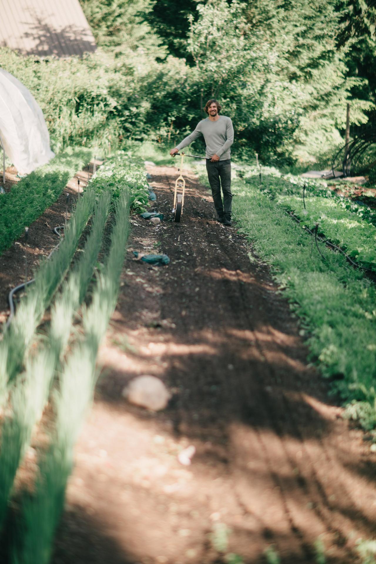 Man in gray long-sleeve shirt holds a single-speed bicycle on a dirt farm path, dense green foliage behind him