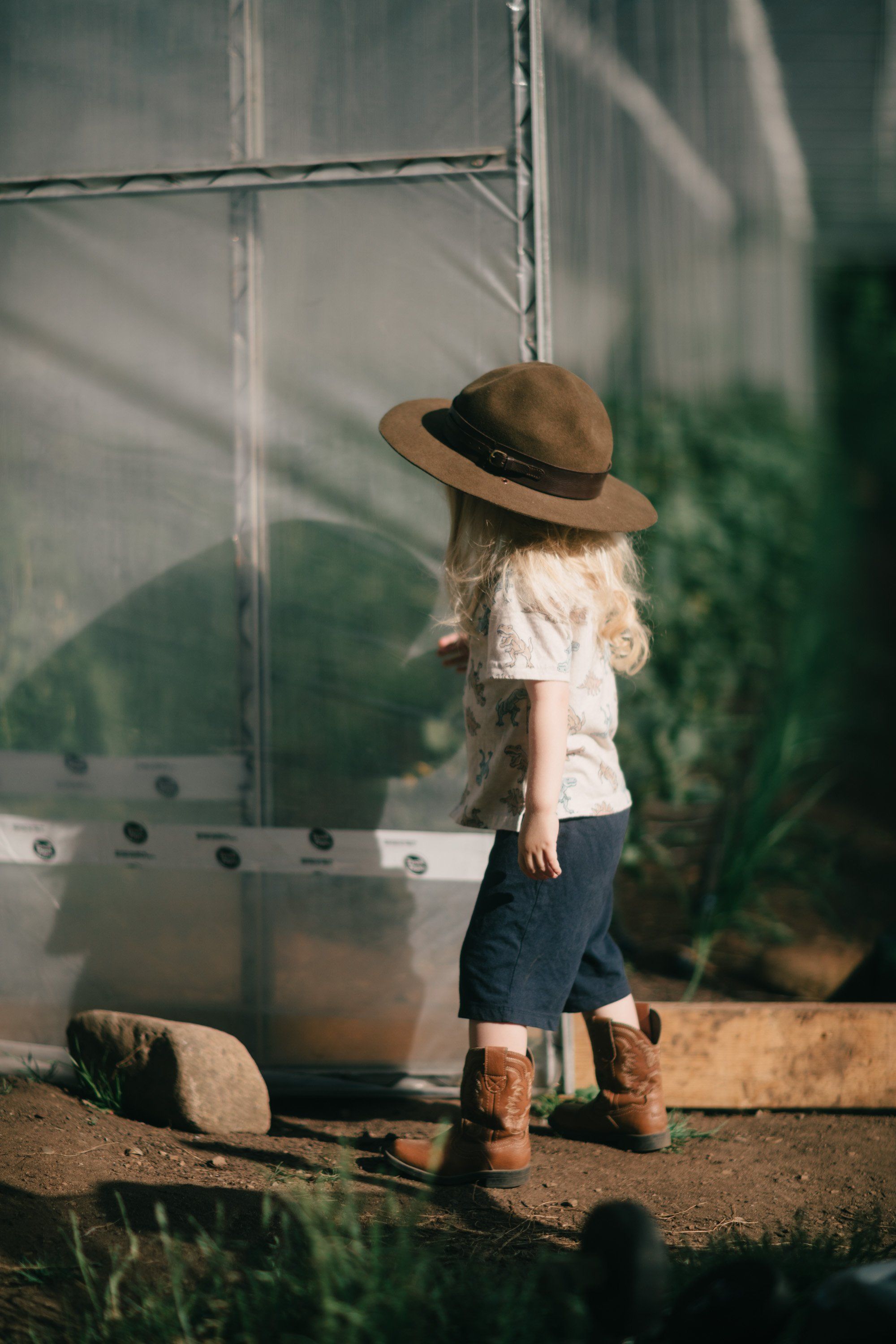 Child in straw hat standing against weathered metal structure with dappled sunlight and green foliage backdrop