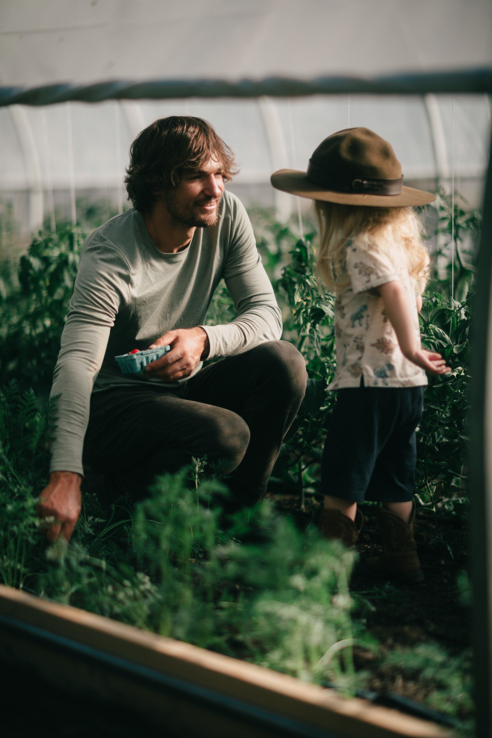Man in cream sweater crouches beside child in hat among lush greenhouse plants, warm afternoon light filtering through white plastic