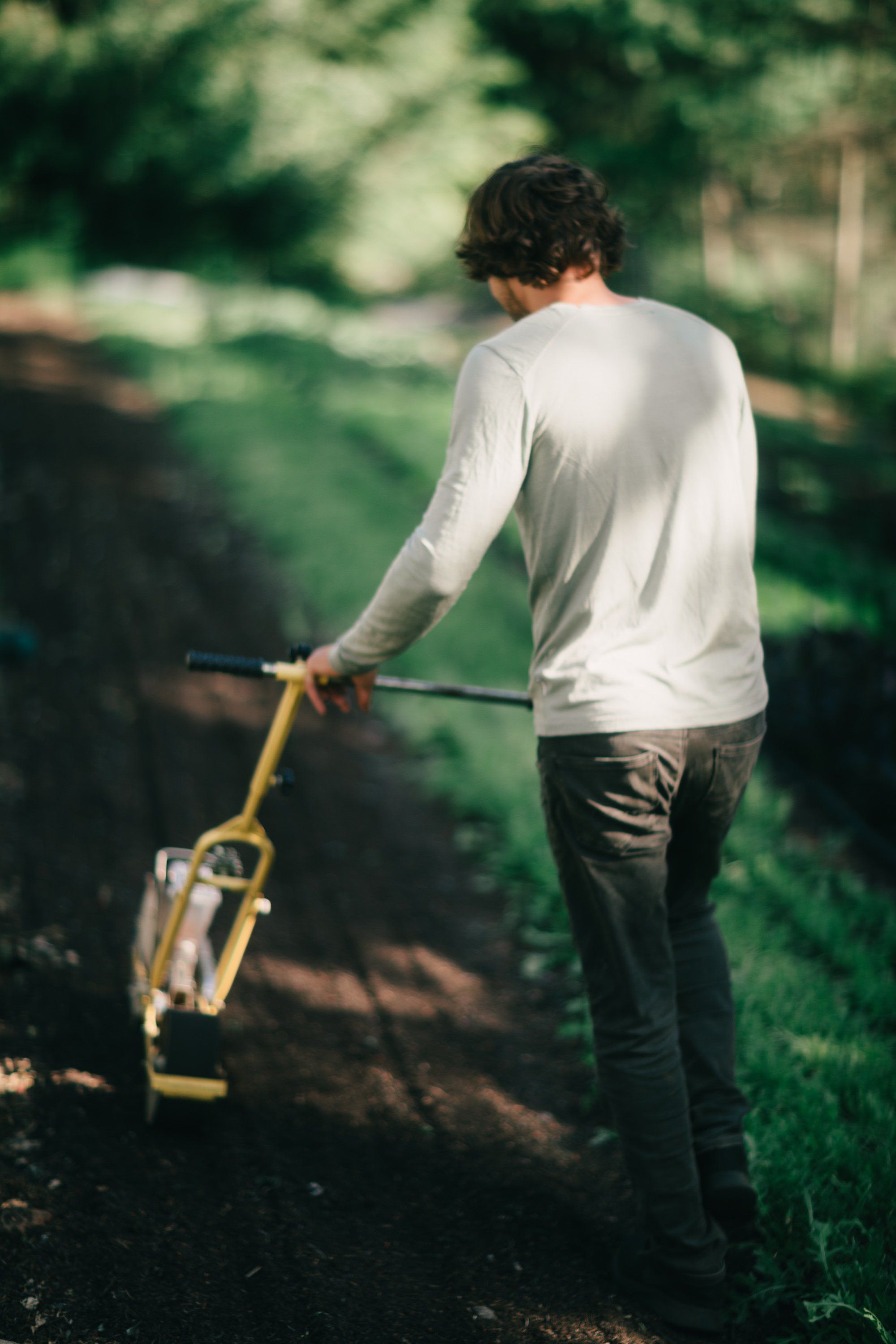 Man in cream long-sleeve shirt holds shovel over garden soil with lush green foliage backlit in afternoon light