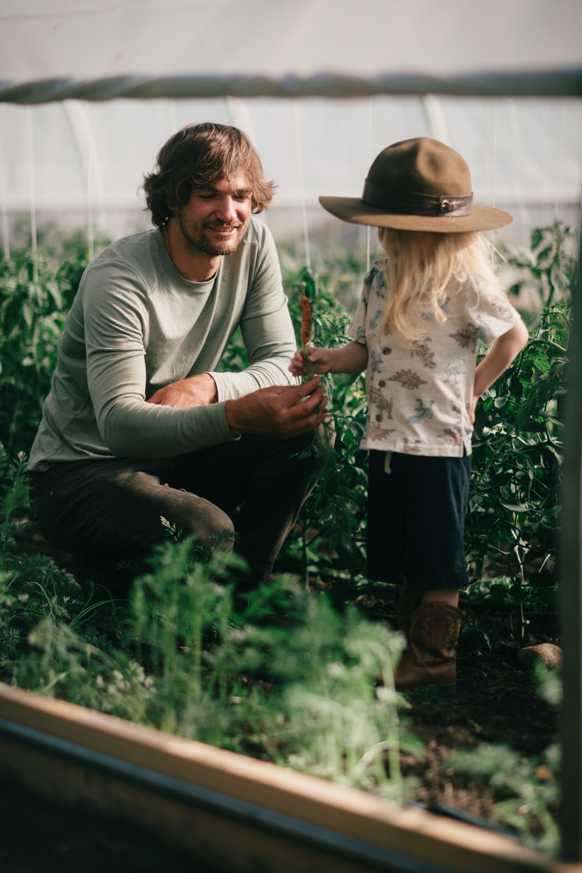 Man crouching beside young child in lush greenhouse garden, soft natural light filtering through white fabric panels