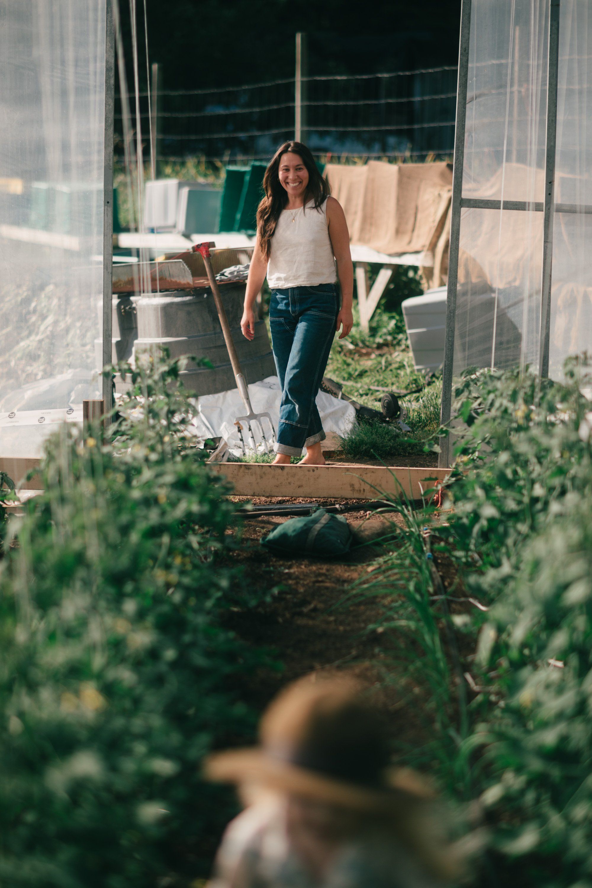 Woman standing in sunlit greenhouse among tomato plants, holding a shovel, with natural light streaming through plastic panels