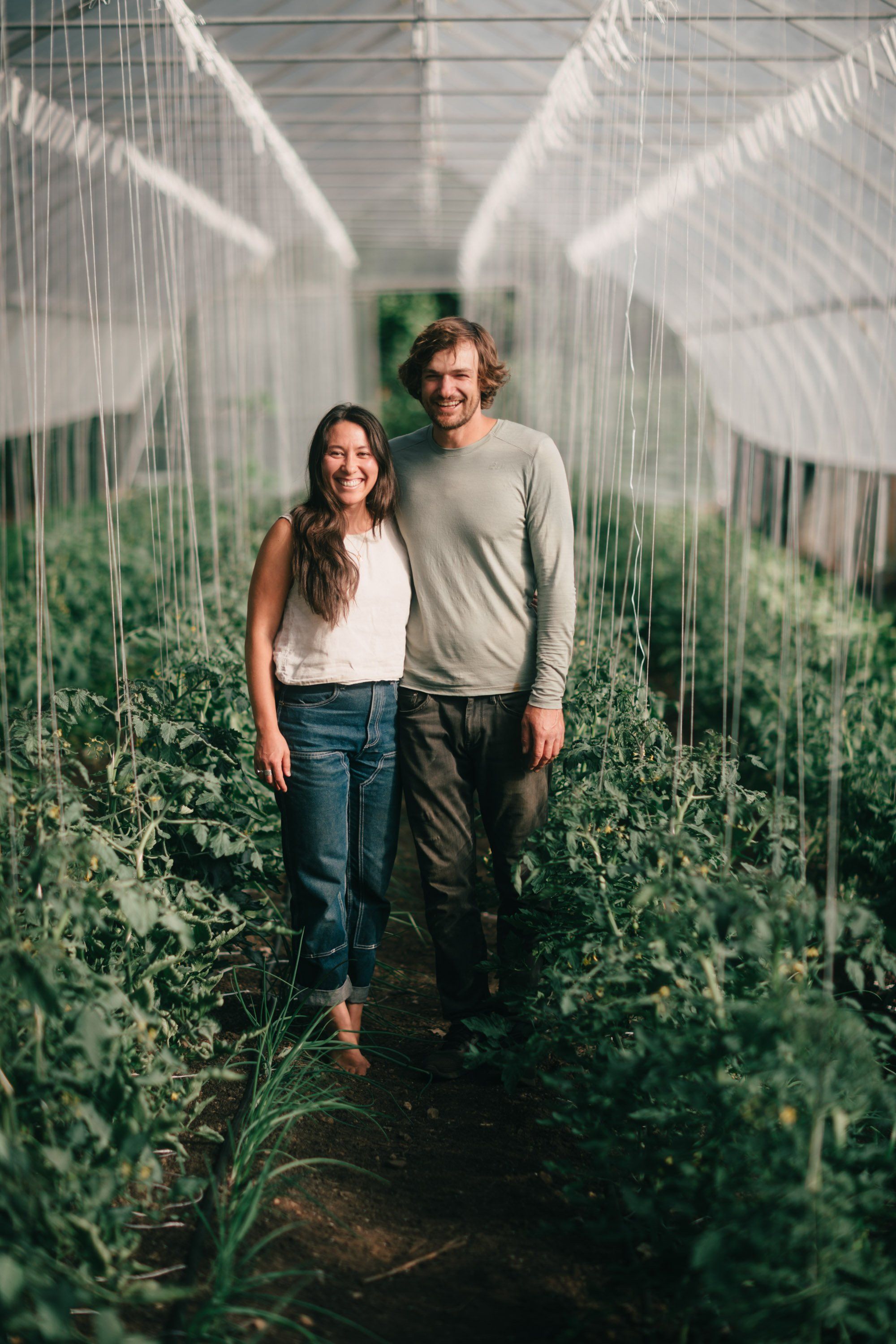 Couple walking hand-in-hand down a lush greenhouse row with white mesh netting overhead, dappled sunlight filtering through the plants
