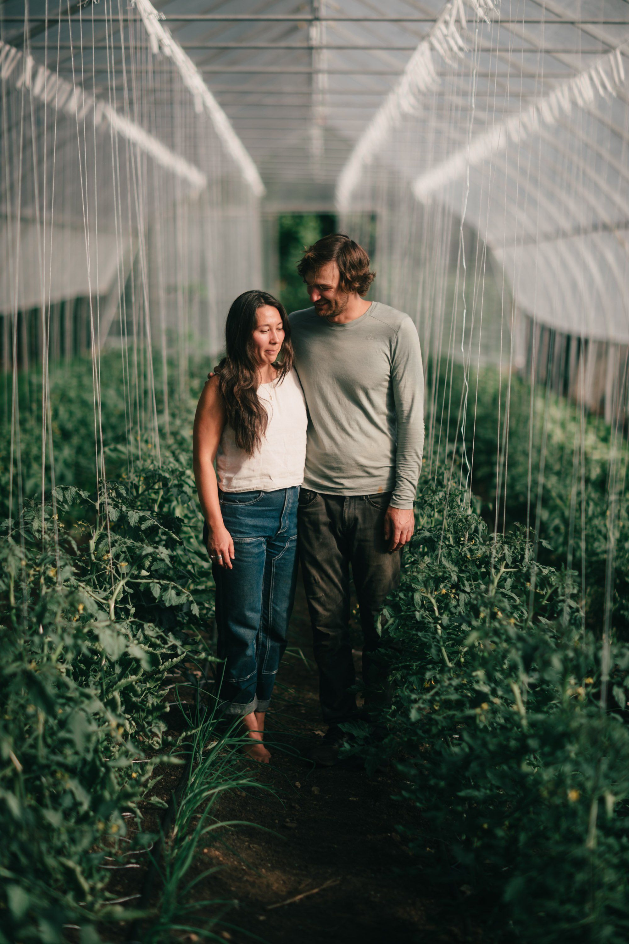 Couple walking through a sunlit greenhouse corridor lined with green plants, dappled shadows across their faces and the white netting overhead