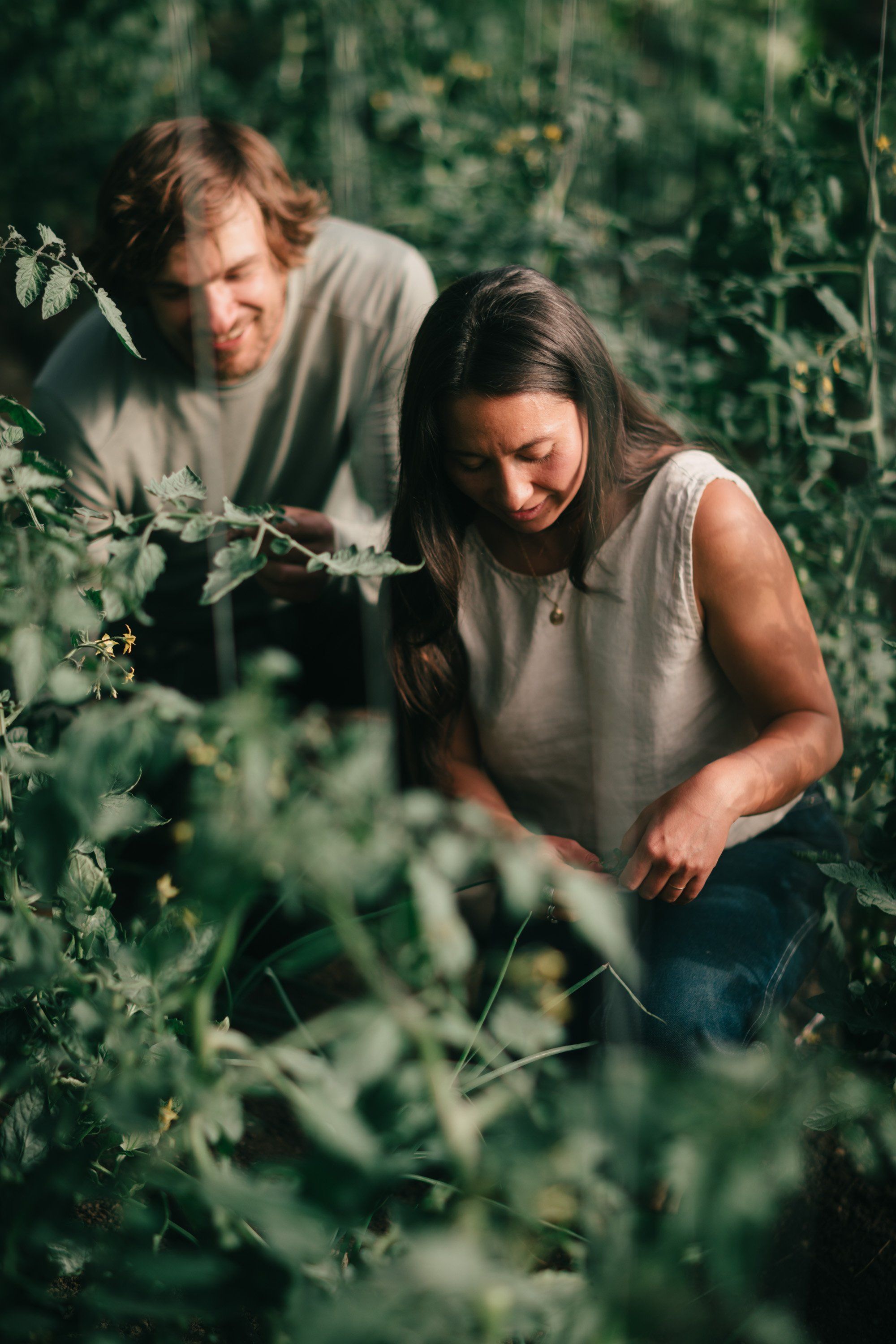Two people in neutral tones explore lush green foliage in dappled natural light, surrounded by wild garden growth